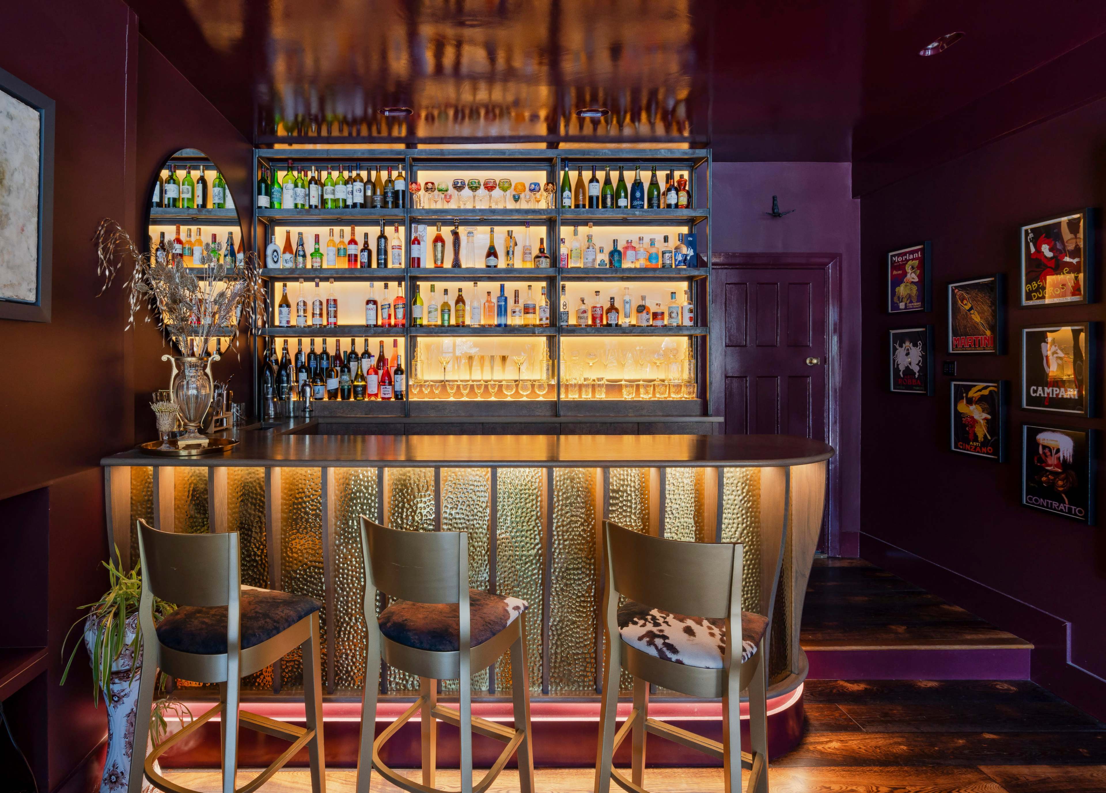 A stylish bar area with a curvilinear counter, shelves lined with various bottles, and three upholstered stools in front.
