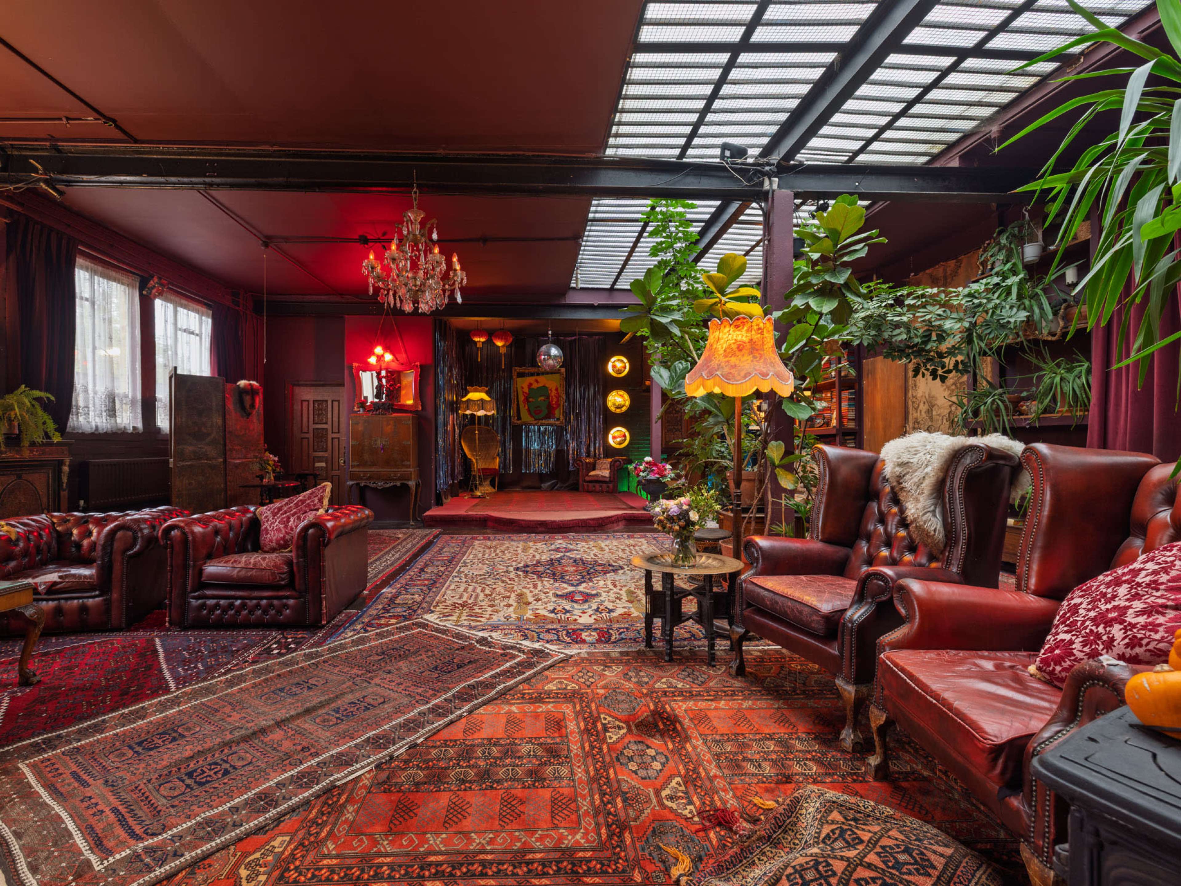 The room features vintage furniture arranged on an ornate patterned rug, with plants and decorative lighting enhancing the eclectic interior.