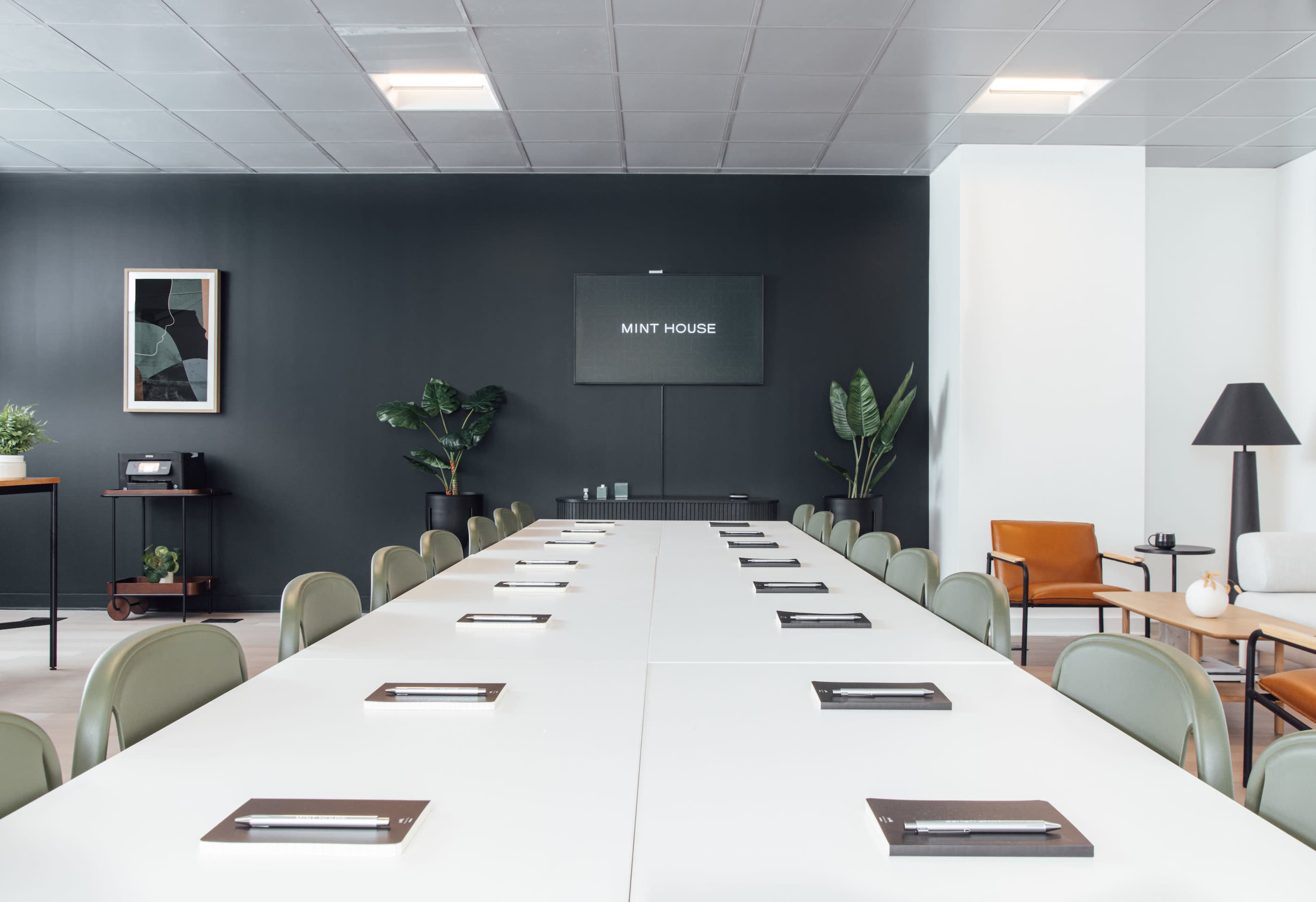 A modern conference room features a long white table with notepads, surrounded by green chairs and set against a dark wall displaying a sign that reads "MINT HOUSE."