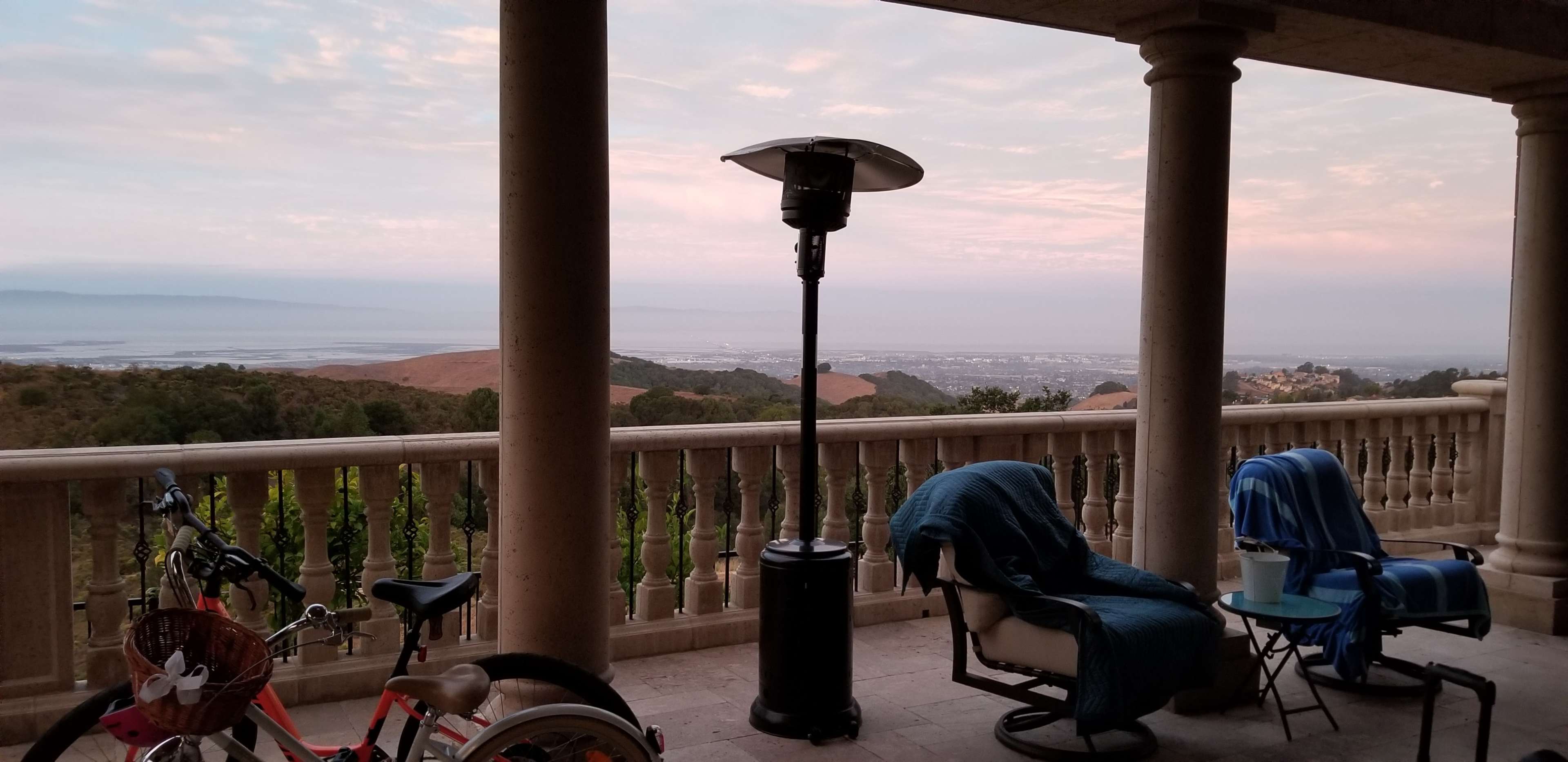 The image shows a covered patio with a heater, two chairs, a small table, and a bicycle, overlooking a hilly landscape and distant water.