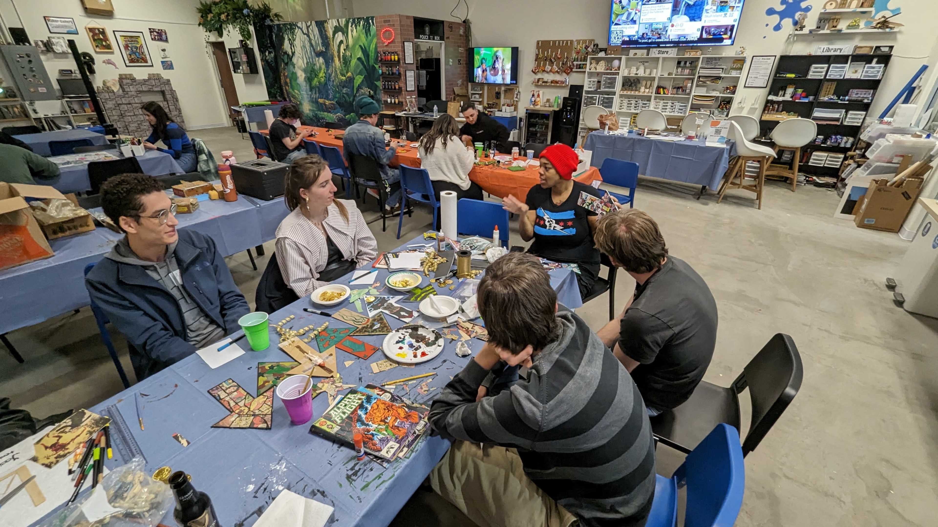 A large group of people is gathered around tables in a bright, open room, engaged in crafting activities while food and drinks are placed nearby.