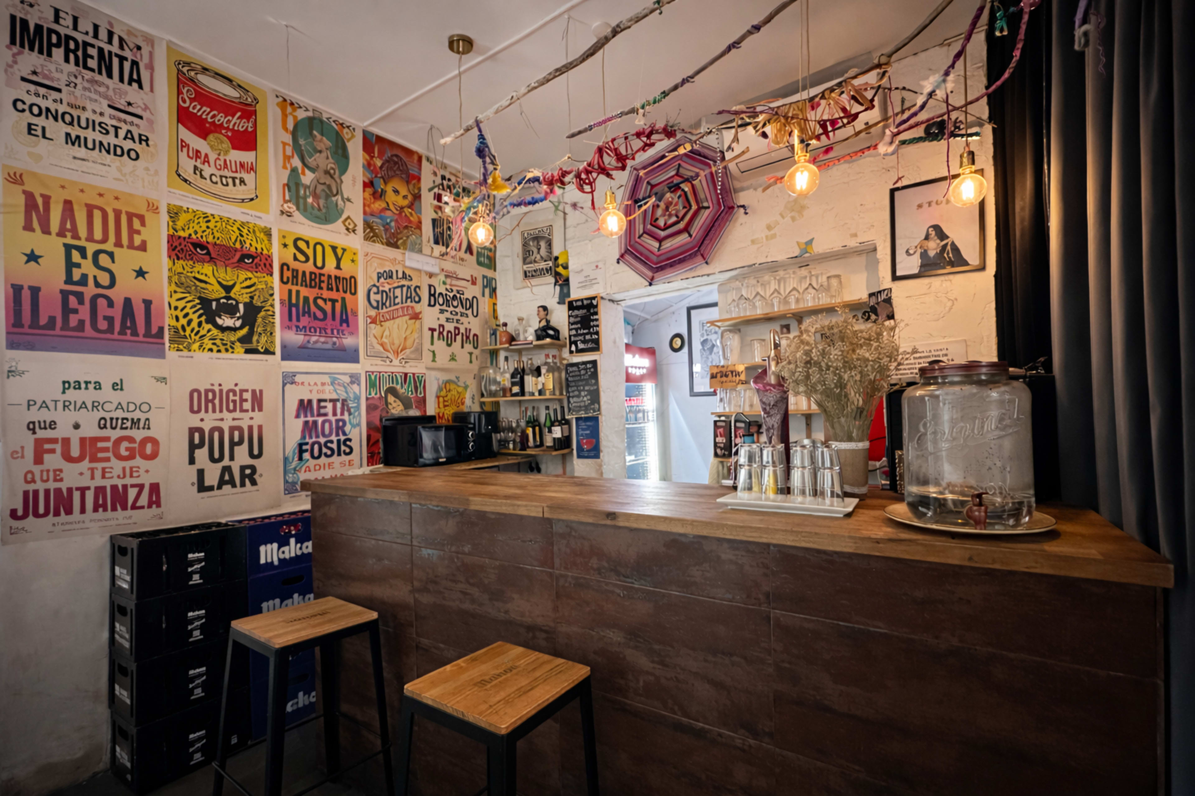 The image shows a lively bar interior with colorful posters on the walls, a wooden counter, and a few stools in front of it.