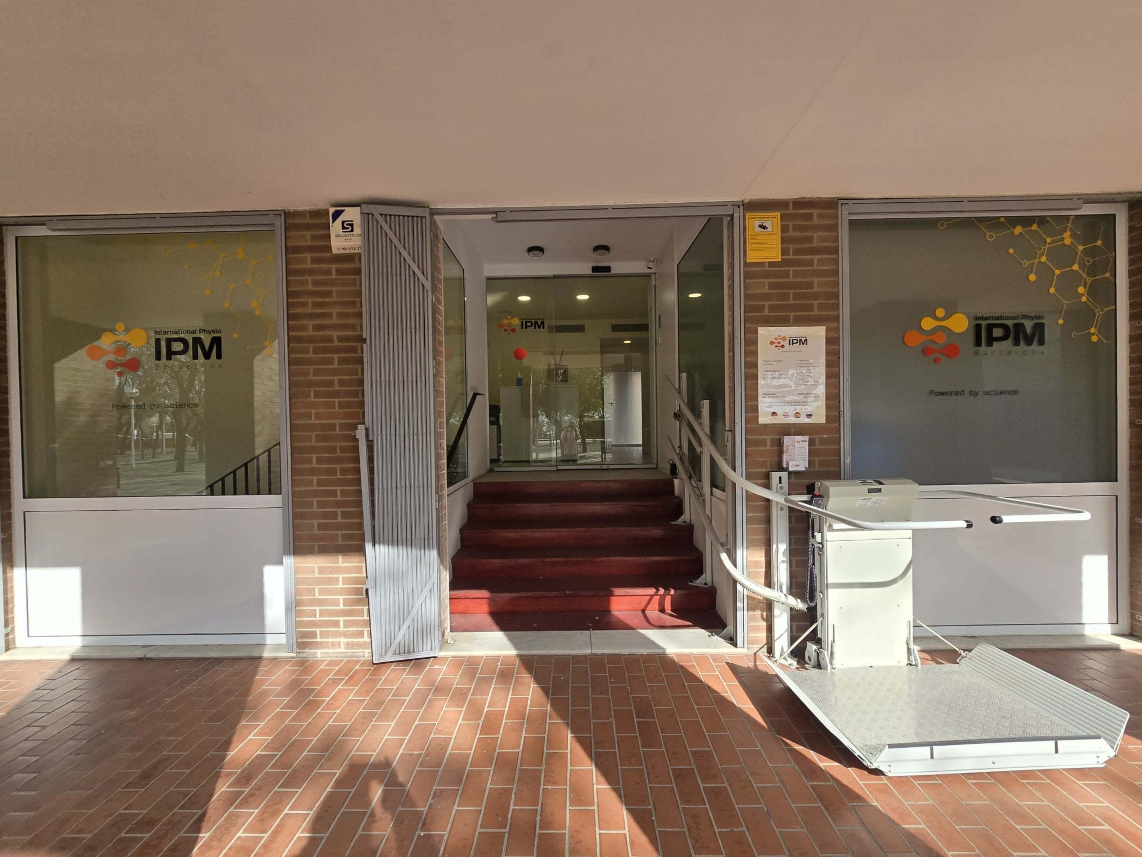 The image shows the entrance of the IPM building with glass doors and a wheelchair ramp leading up to the red carpeted steps.