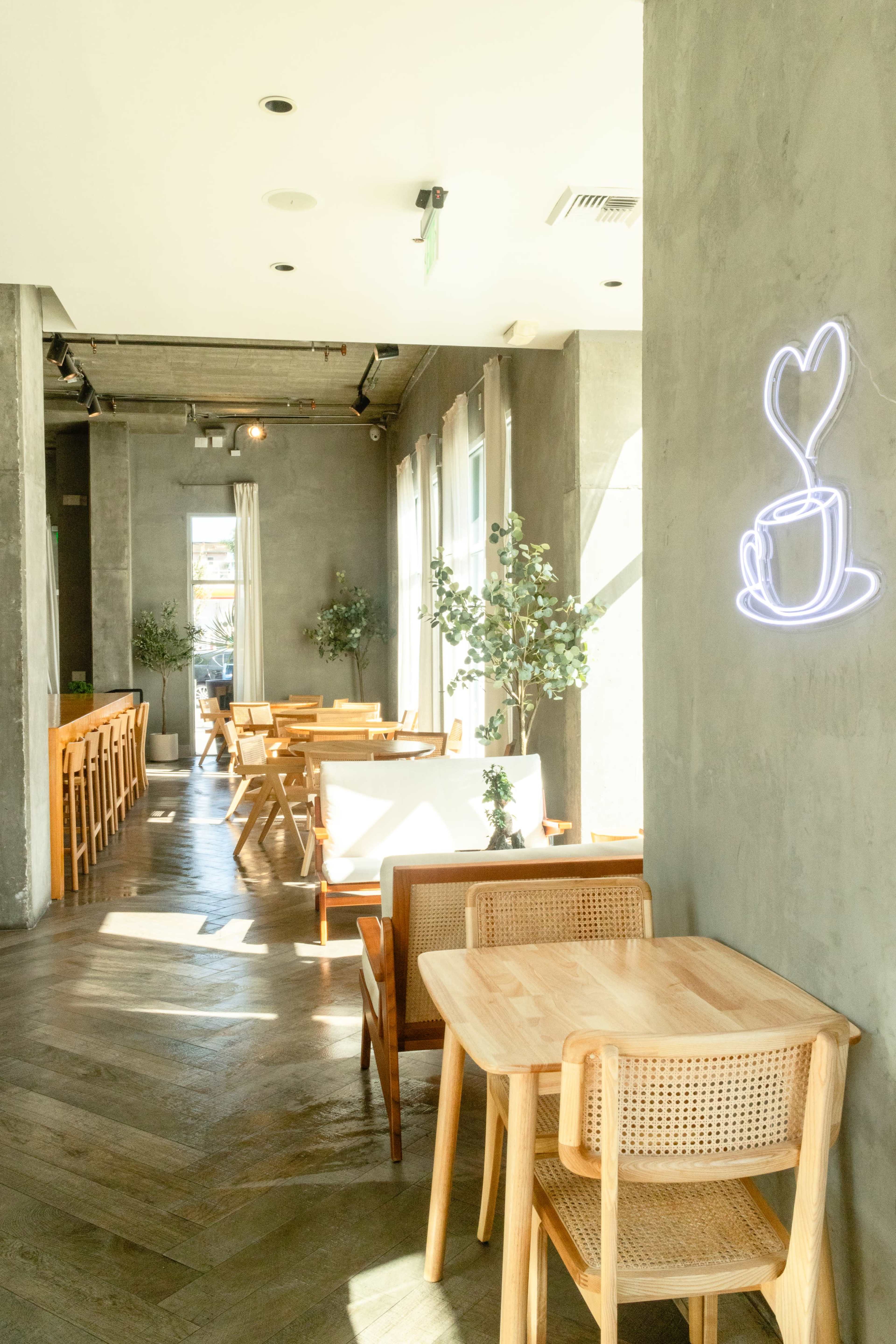 A modern café interior with wooden tables and chairs, large windows, and a neon sign depicting a heart and a cup of coffee.