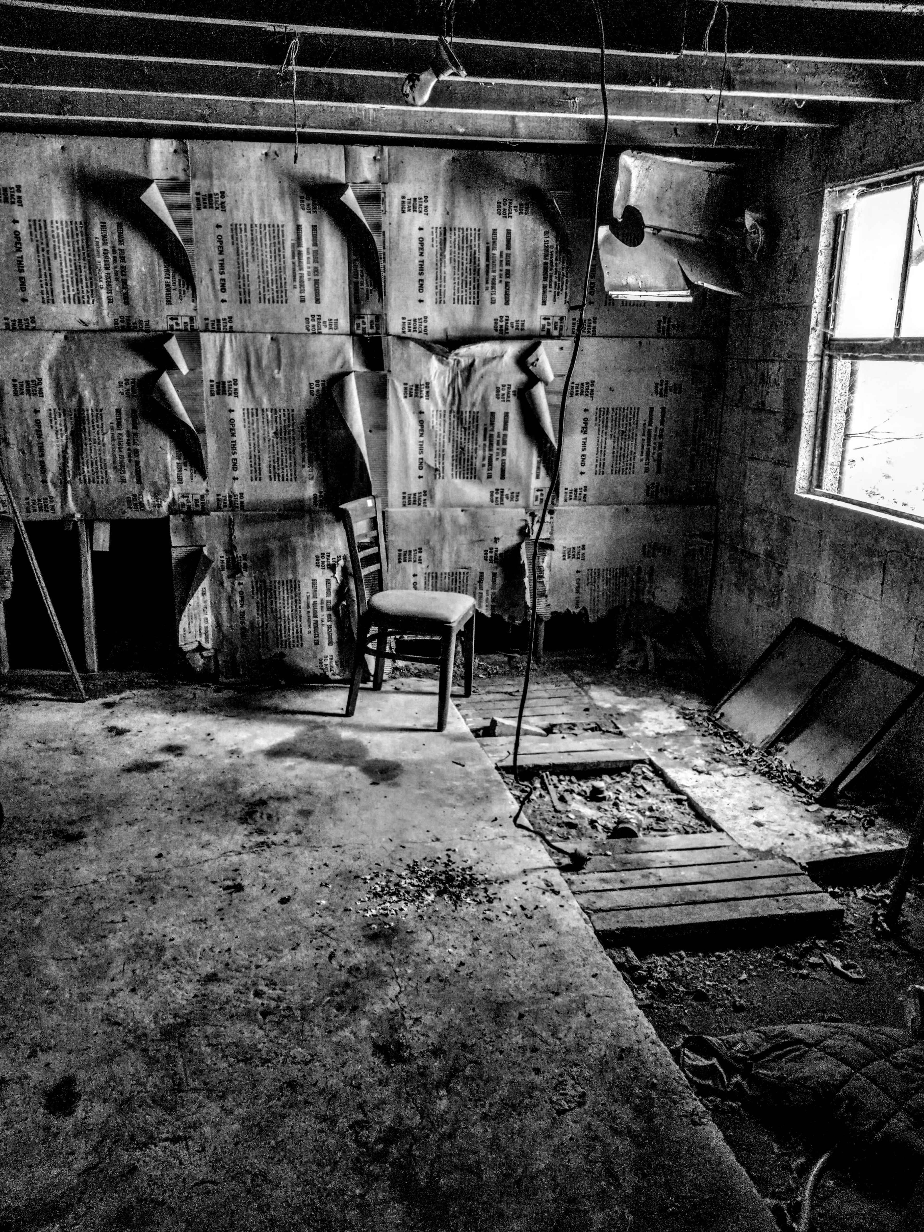 A single wooden chair stands in a dimly lit, abandoned room with bare concrete walls and scattered debris on the floor.