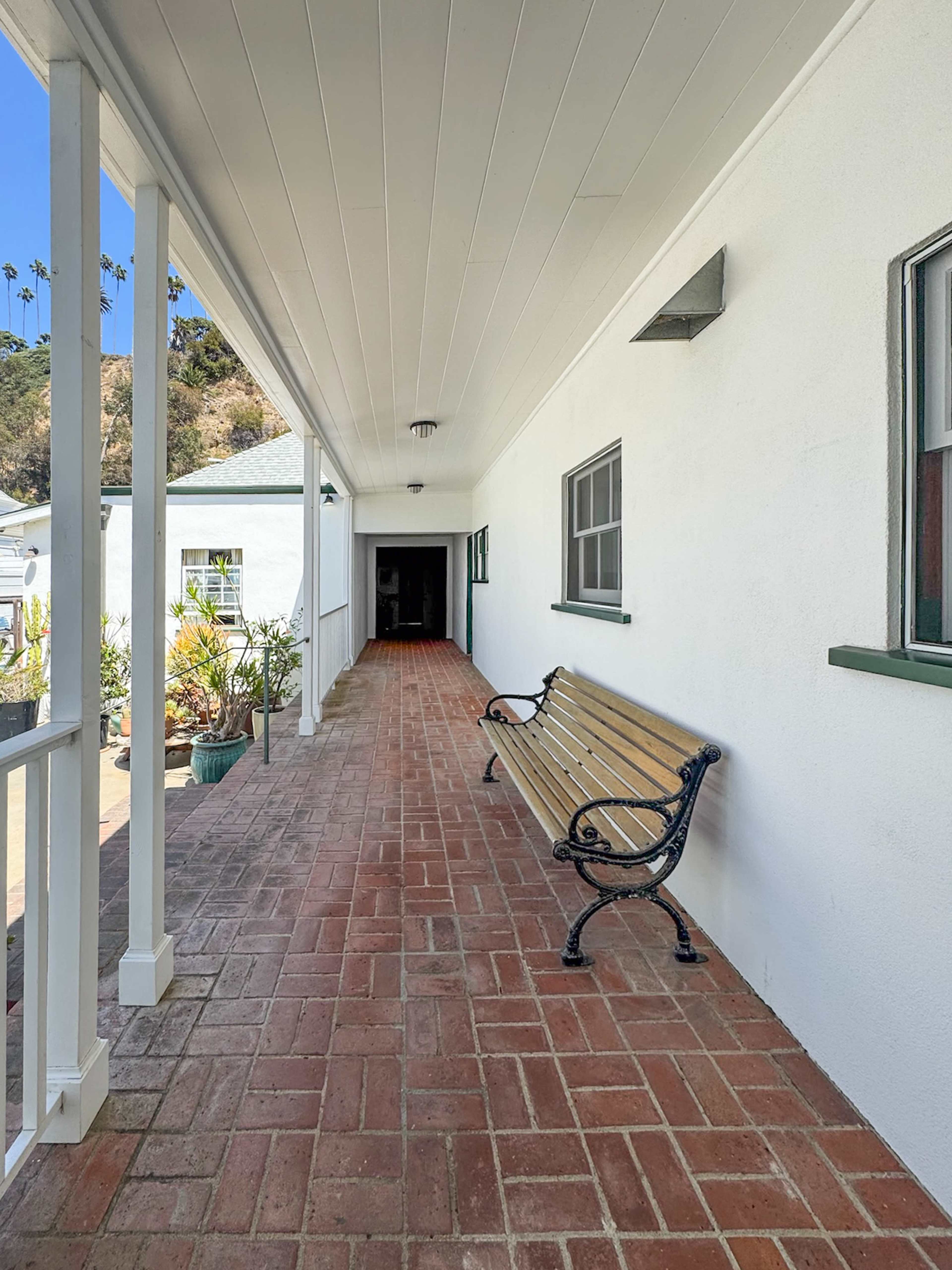 The image shows a long, narrow porch lined with brick flooring and a wooden bench, leading to a doorway at the far end.