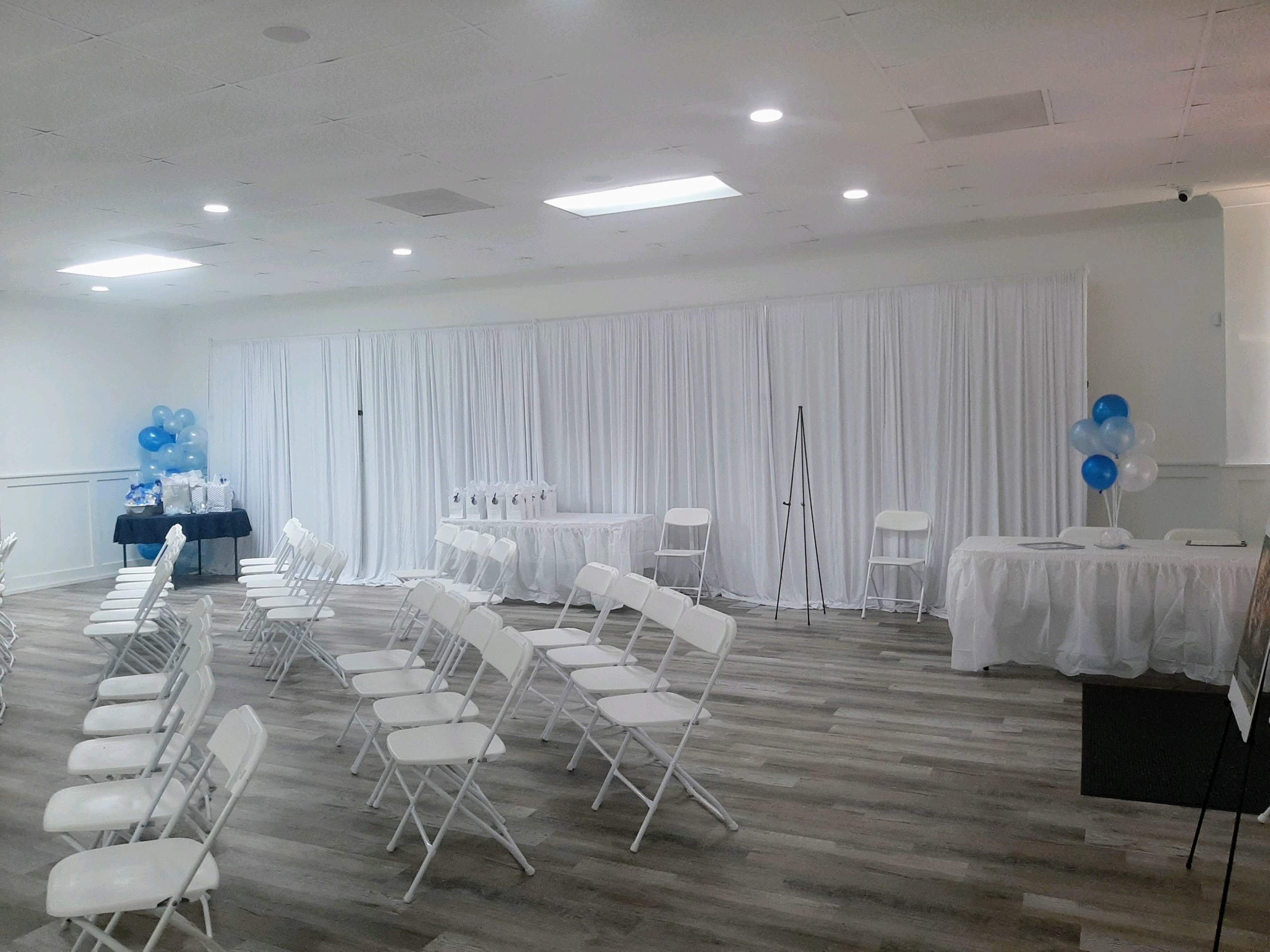 A spacious event room is set up with rows of white chairs facing a display area featuring tables draped in white and blue, along with balloons in the same colors.