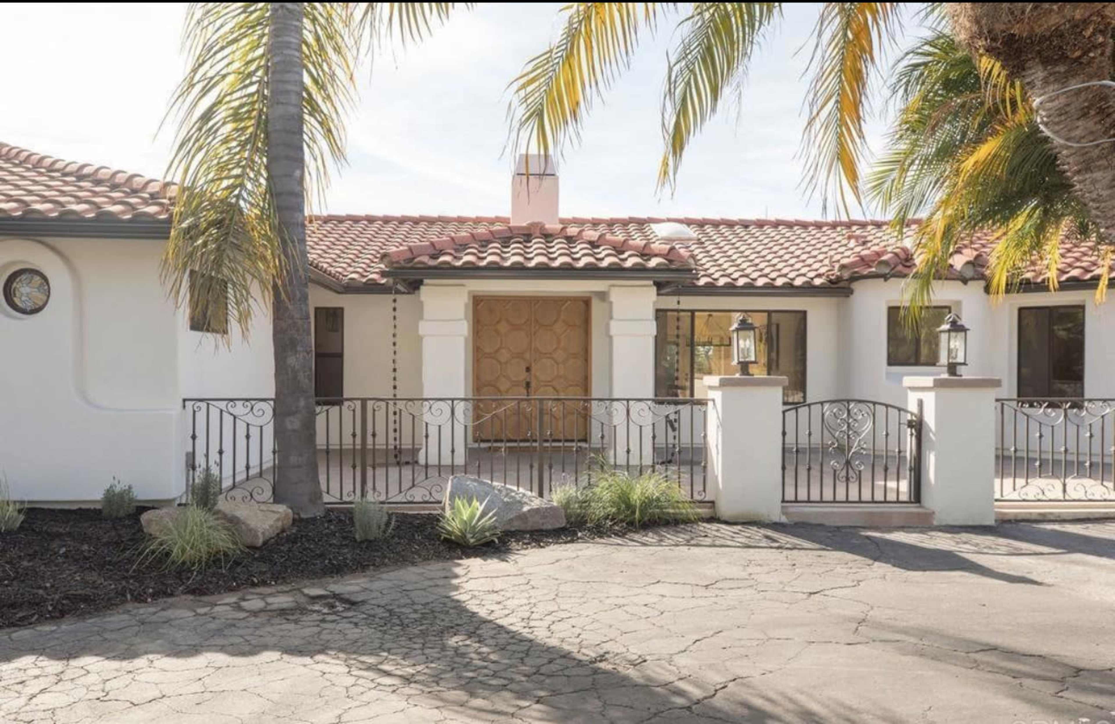 The image shows a Spanish-style house with a terra cotta tile roof, a large wooden front door, and a decorative wrought iron railing in the front courtyard, surrounded by palm trees.