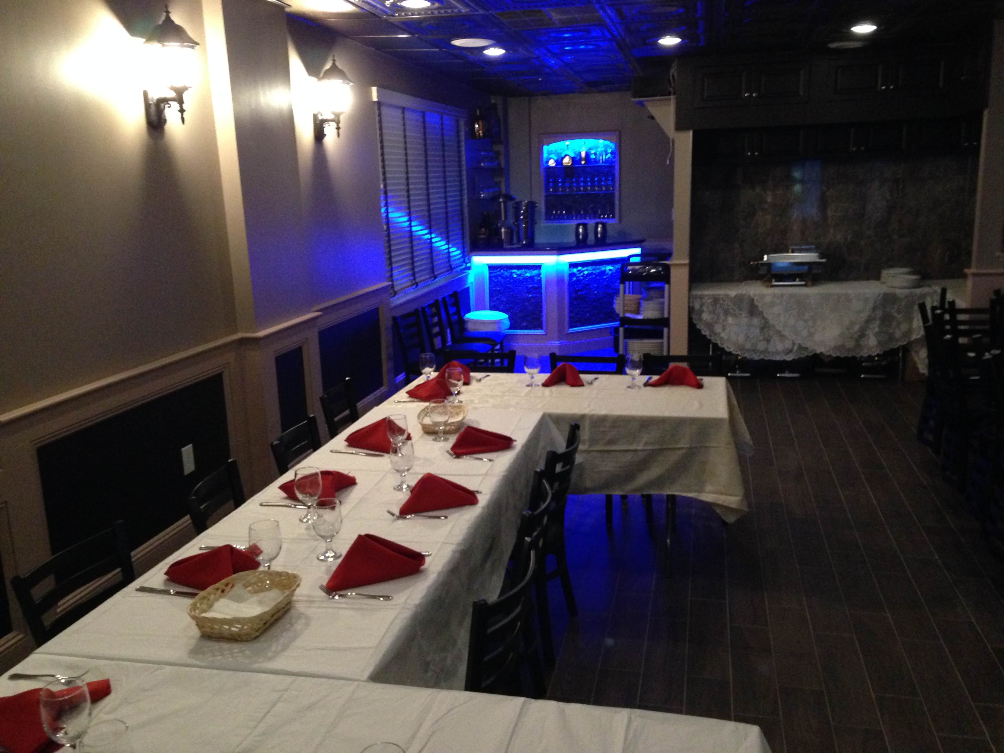 A restaurant dining area with neatly arranged tables covered in white tablecloths and red napkins, featuring a bar area illuminated by blue lighting.