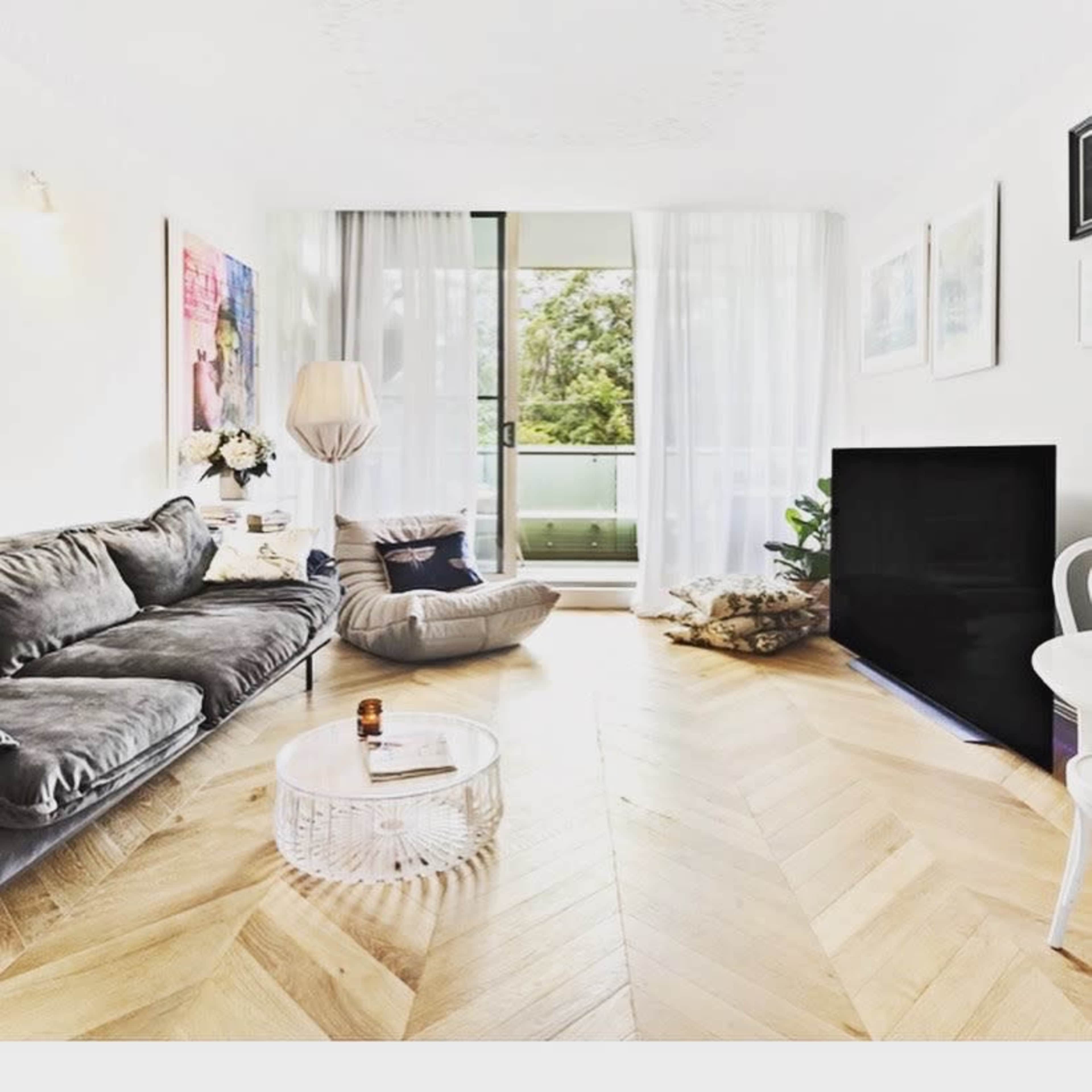 The image shows a modern living room featuring a gray sofa, a cozy armchair, a large television, and hardwood flooring, with natural light streaming in through large windows.