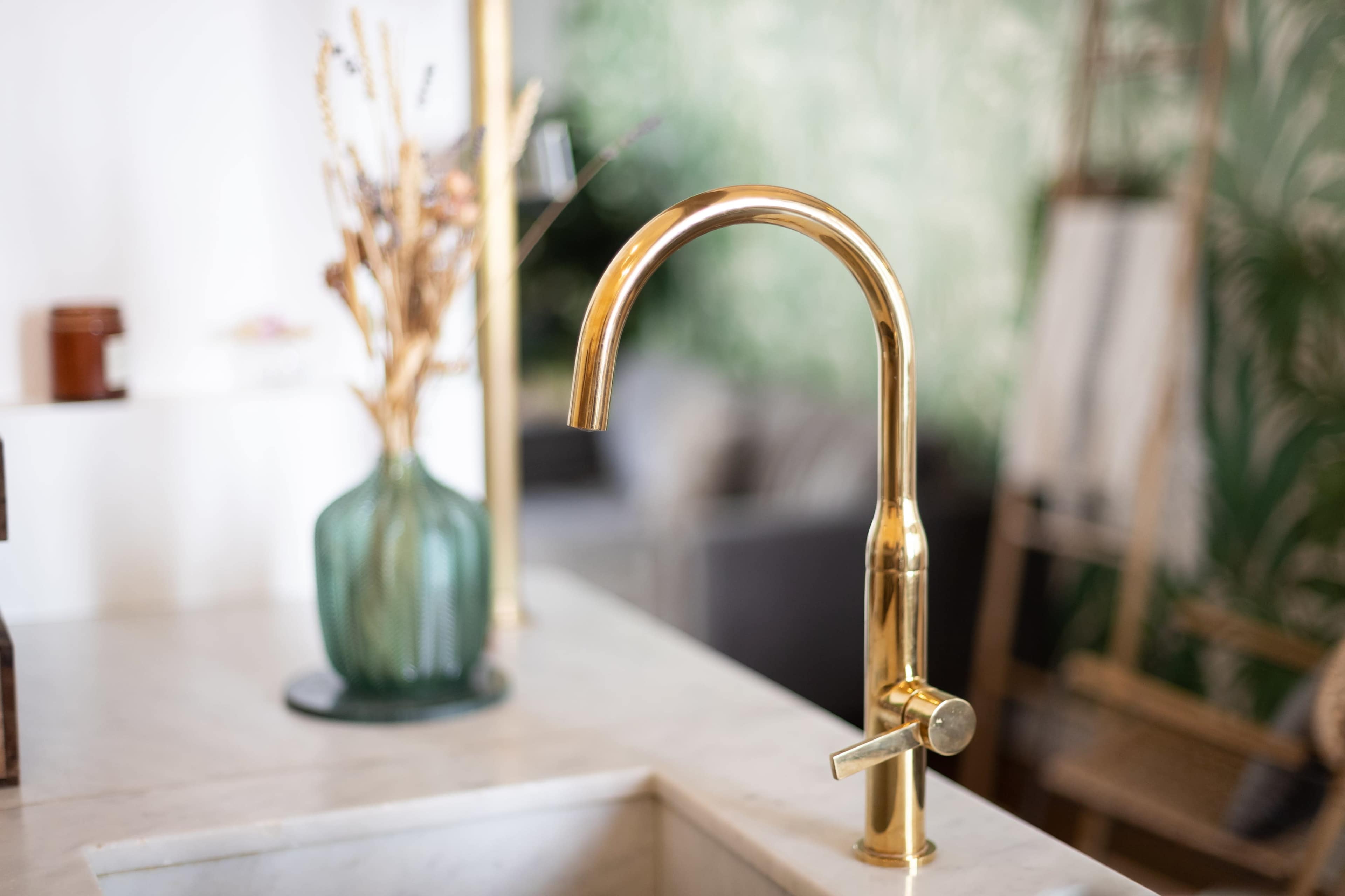 The image shows a modern gold kitchen faucet next to a green glass vase filled with dried flowers on a countertop.