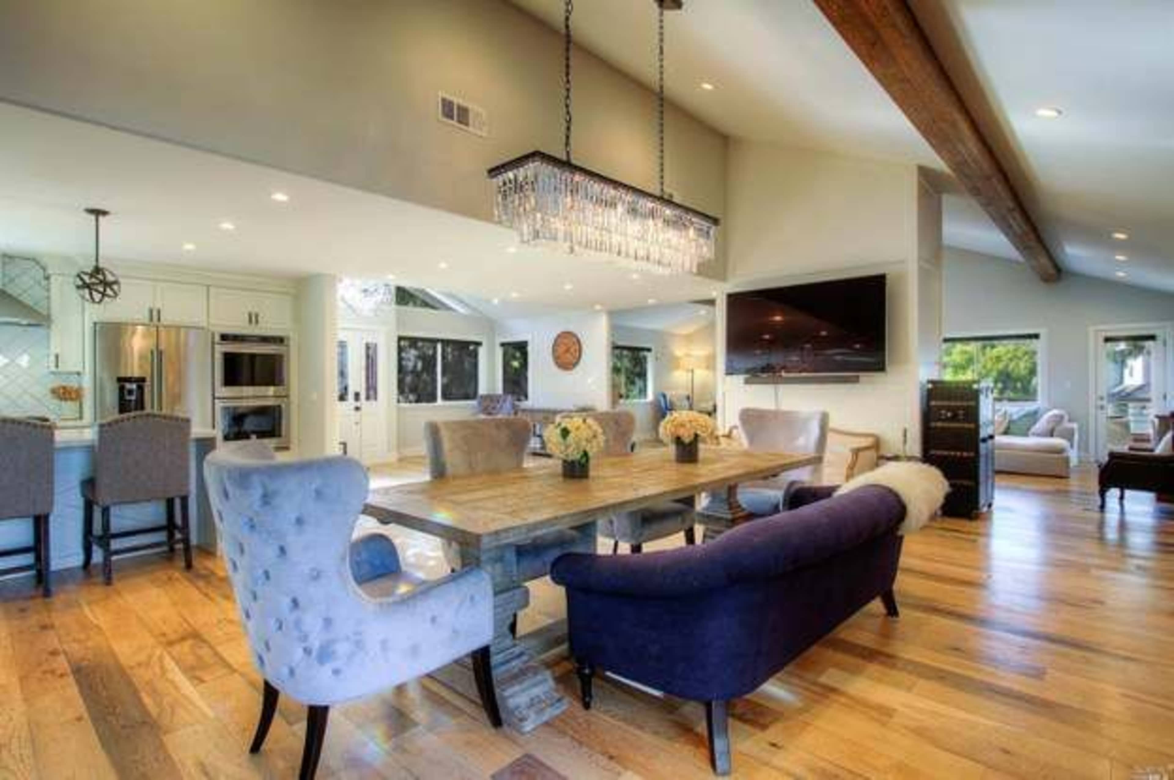 A modern dining area featuring a large wooden table surrounded by upholstered chairs, with a crystal chandelier overhead and a view of a bright kitchen in the background.
