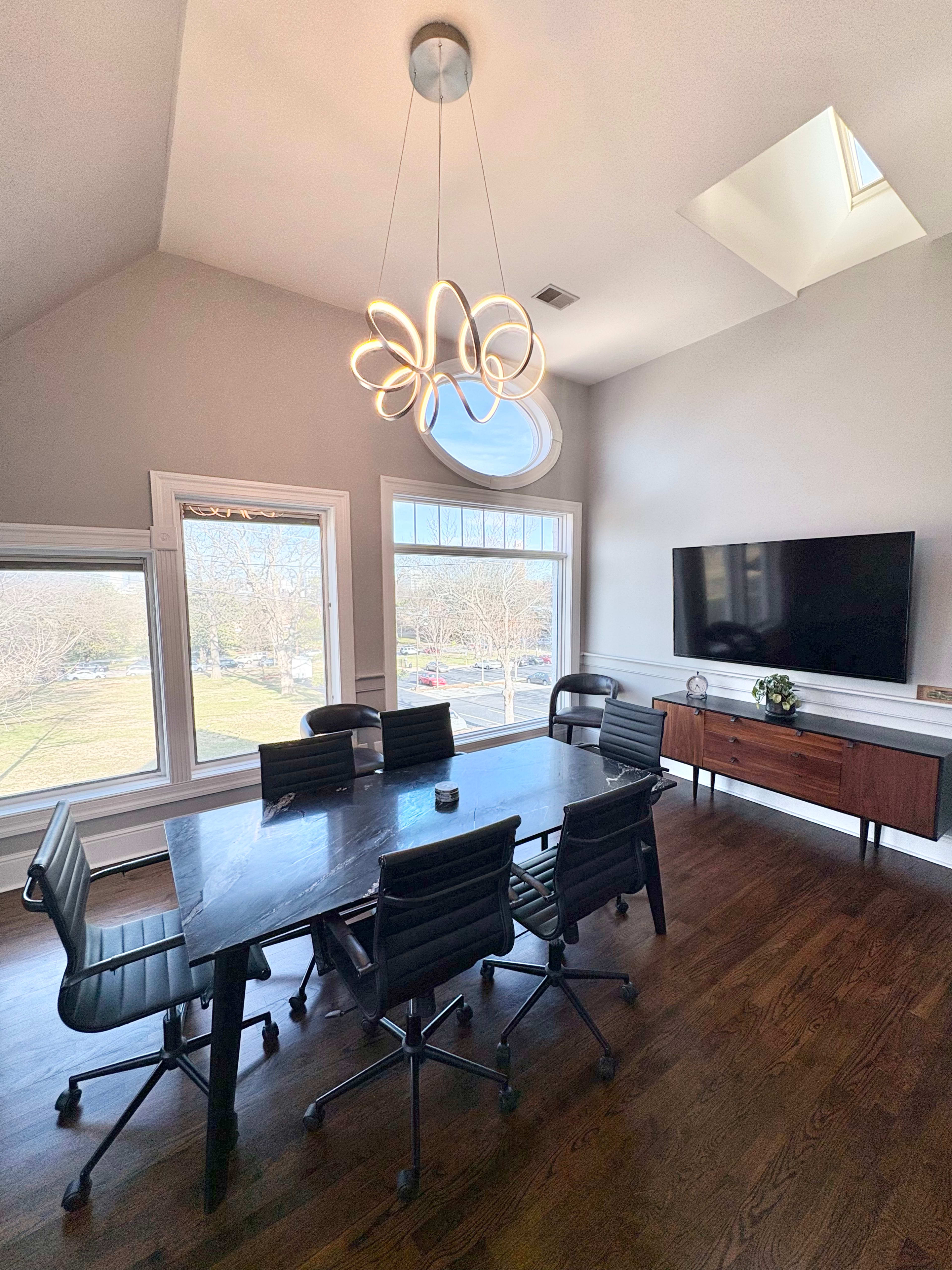 A modern conference room features a large table surrounded by black rolling chairs, with a skylight above and a TV mounted on the wall.