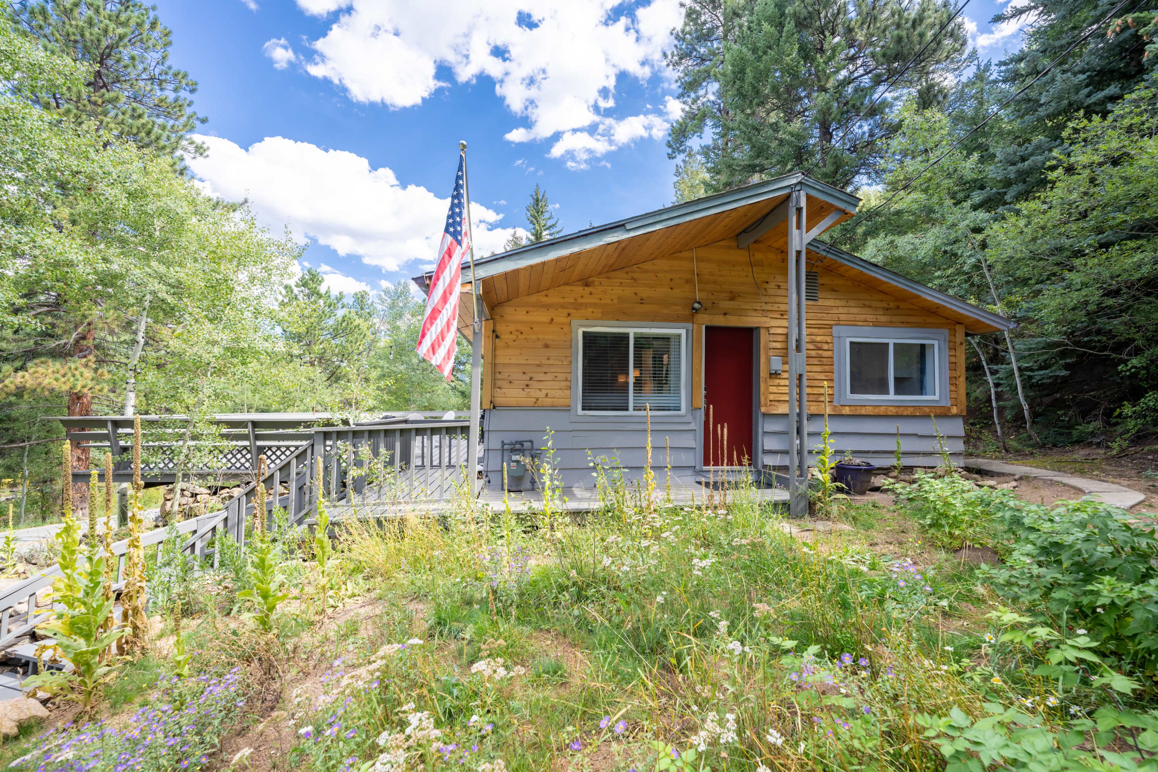 A wooden cabin with an American flag is situated among trees, surrounded by a garden with wildflowers.