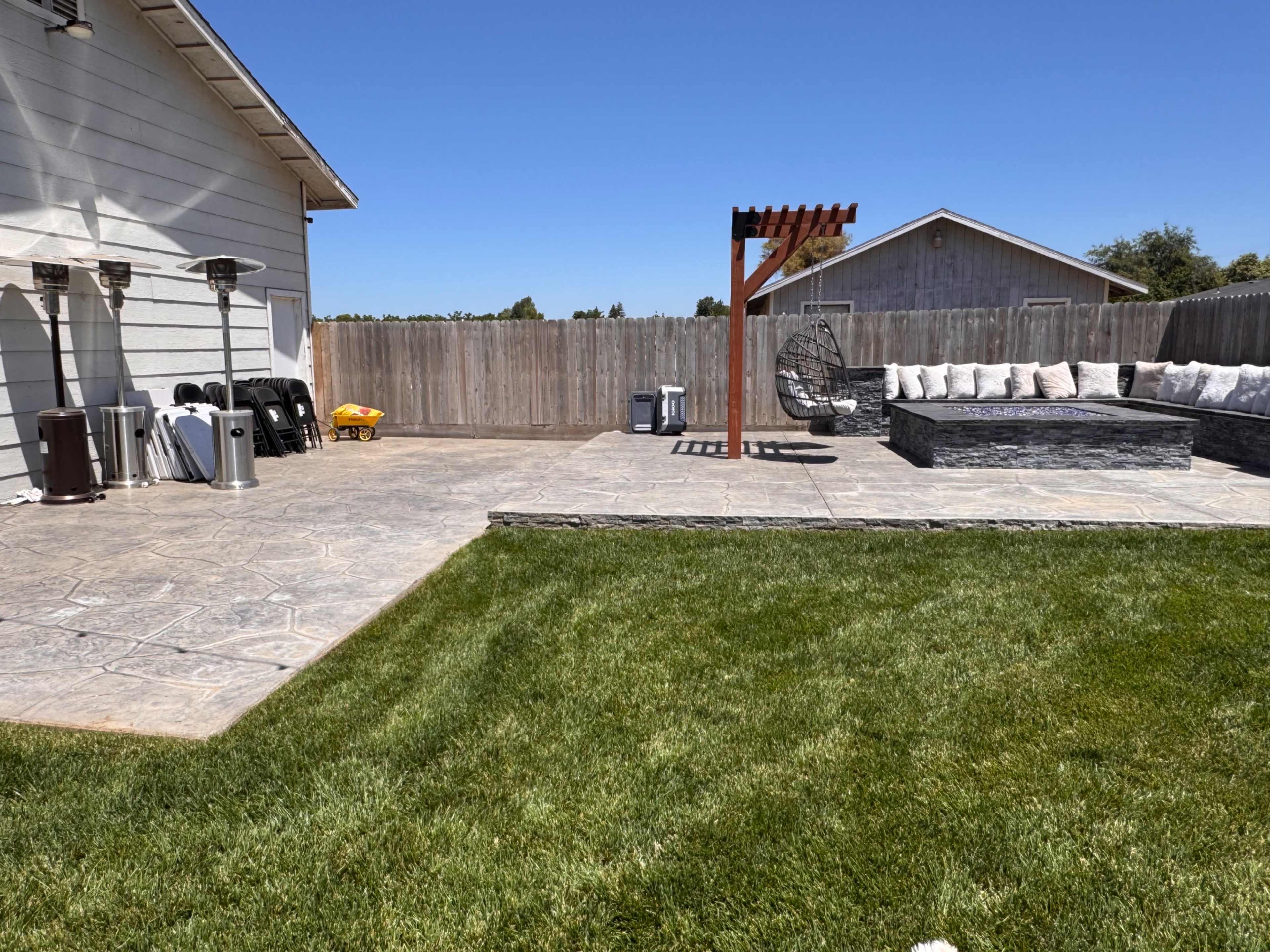 The image shows a backyard with a concrete patio, a grass area, a seating area with cushions, and a wooden pergola.