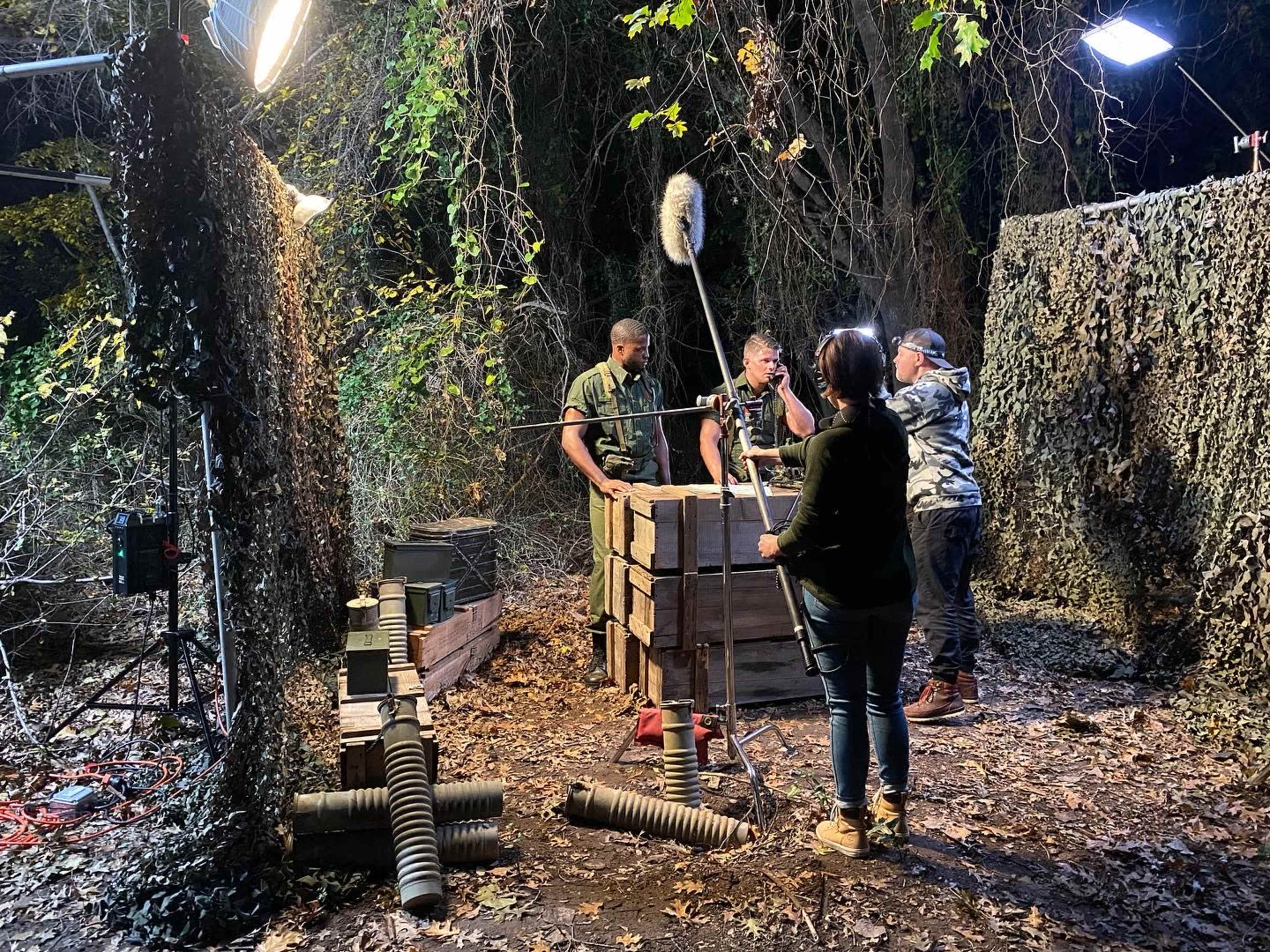 A film crew sets up a scene in a forested area with camouflage backdrops and wooden crates.