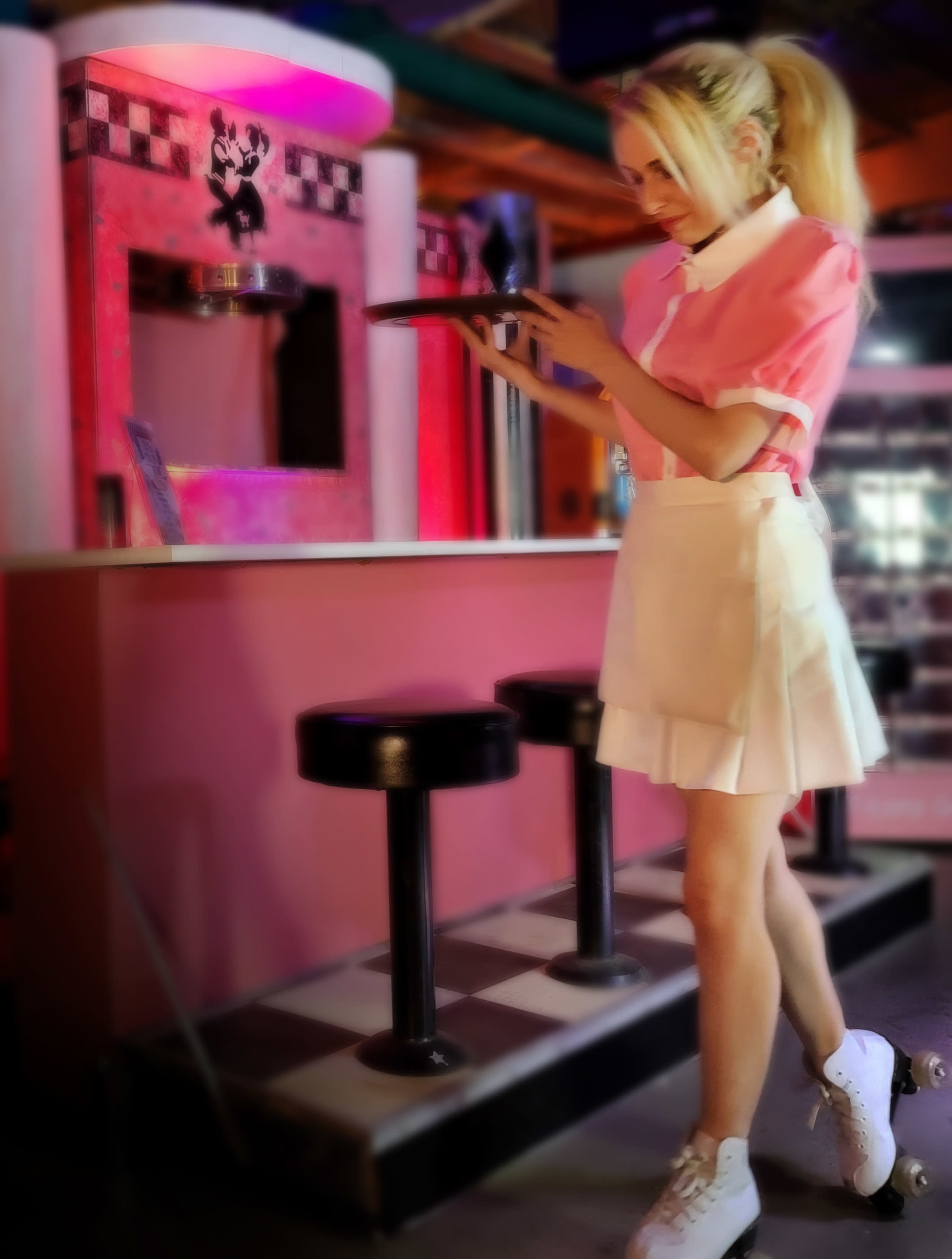 A woman in a pink shirt and white skirt stands at a retro-style counter, holding a tablet while wearing roller skates.