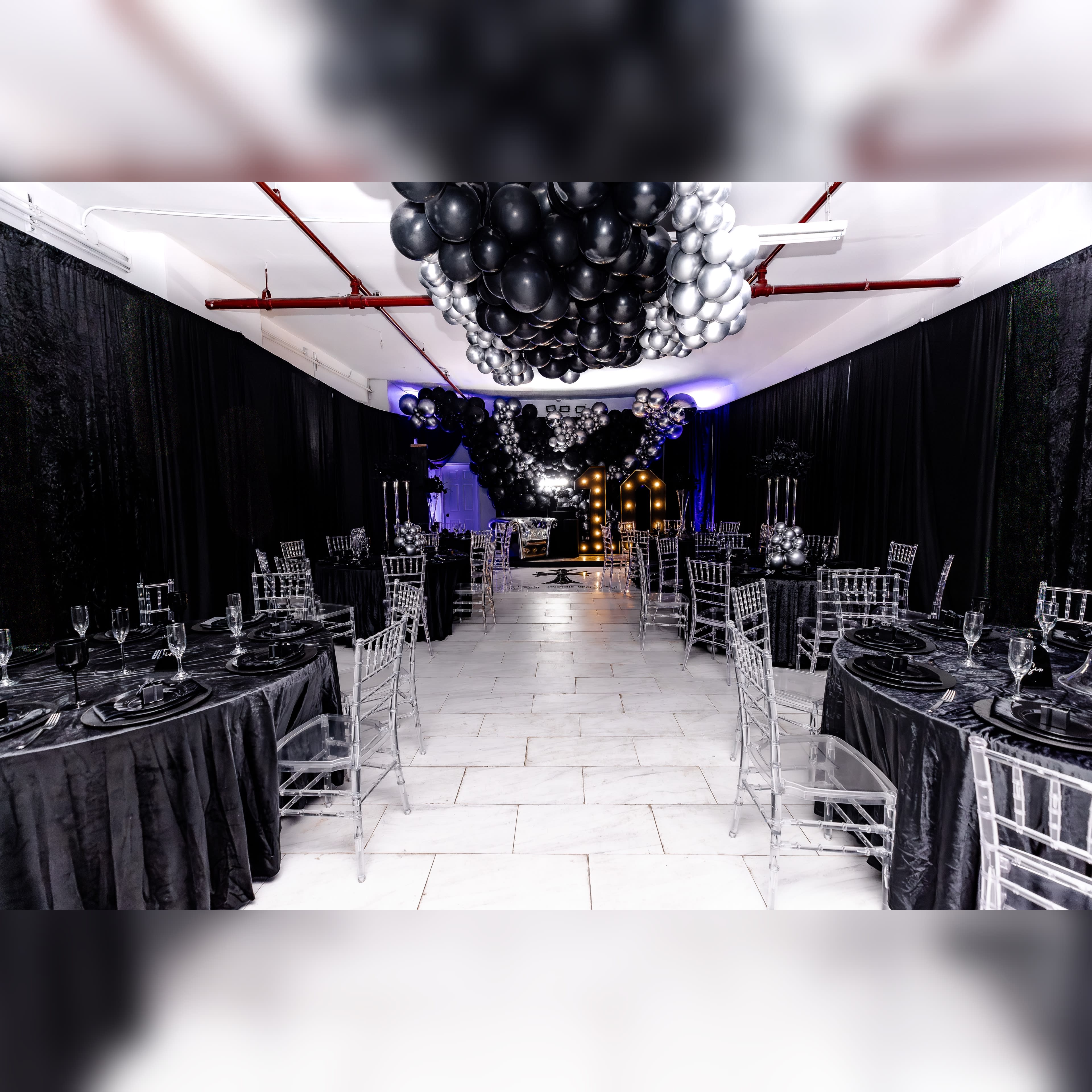 The image shows a decorated event space featuring round tables with black tablecloths and clear chairs, surrounded by black and silver balloon arrangements.