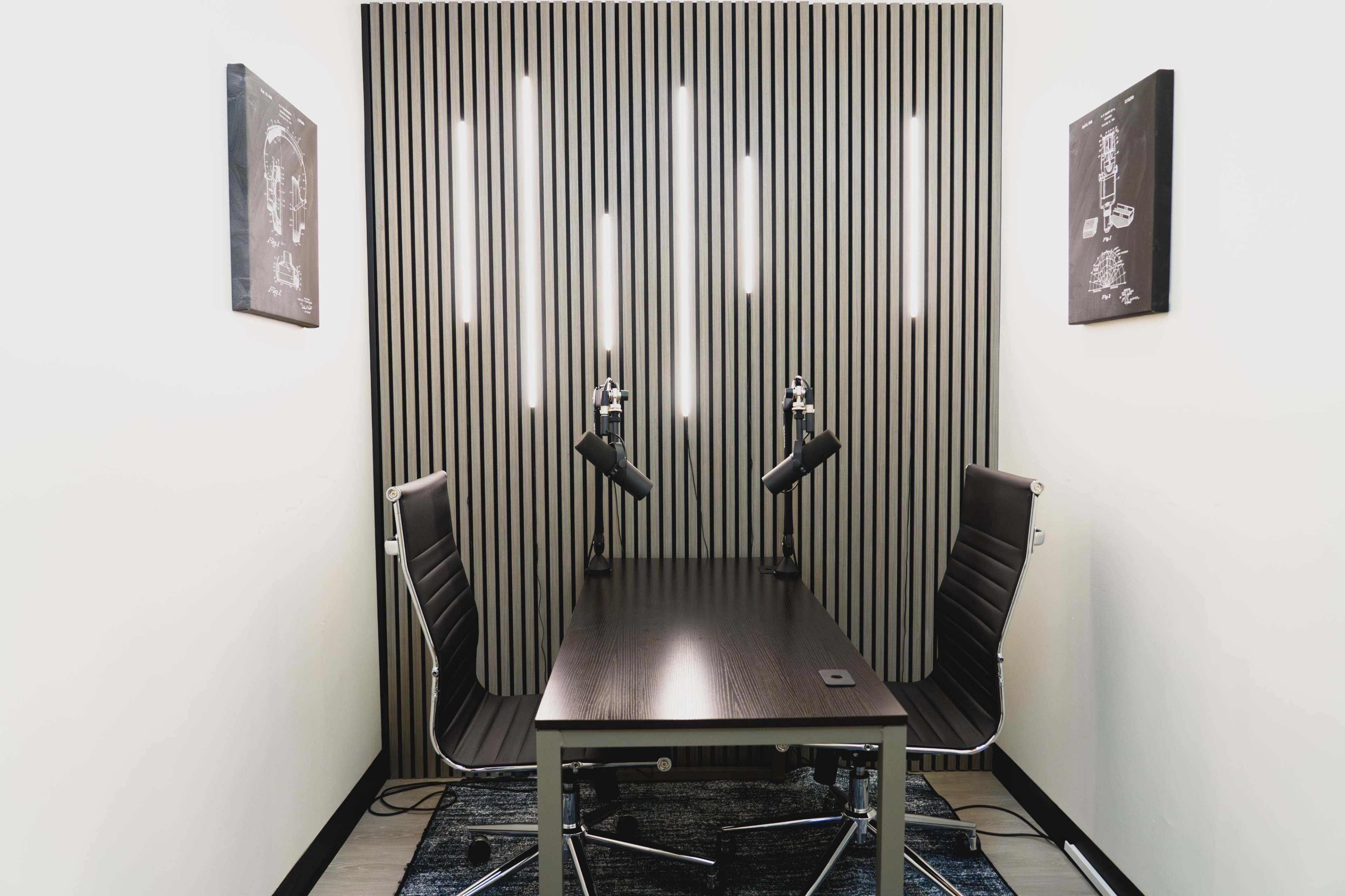The image shows a minimalistic meeting room with two black chairs and a sleek table, accented by wall-mounted lights and framed artwork on the walls.