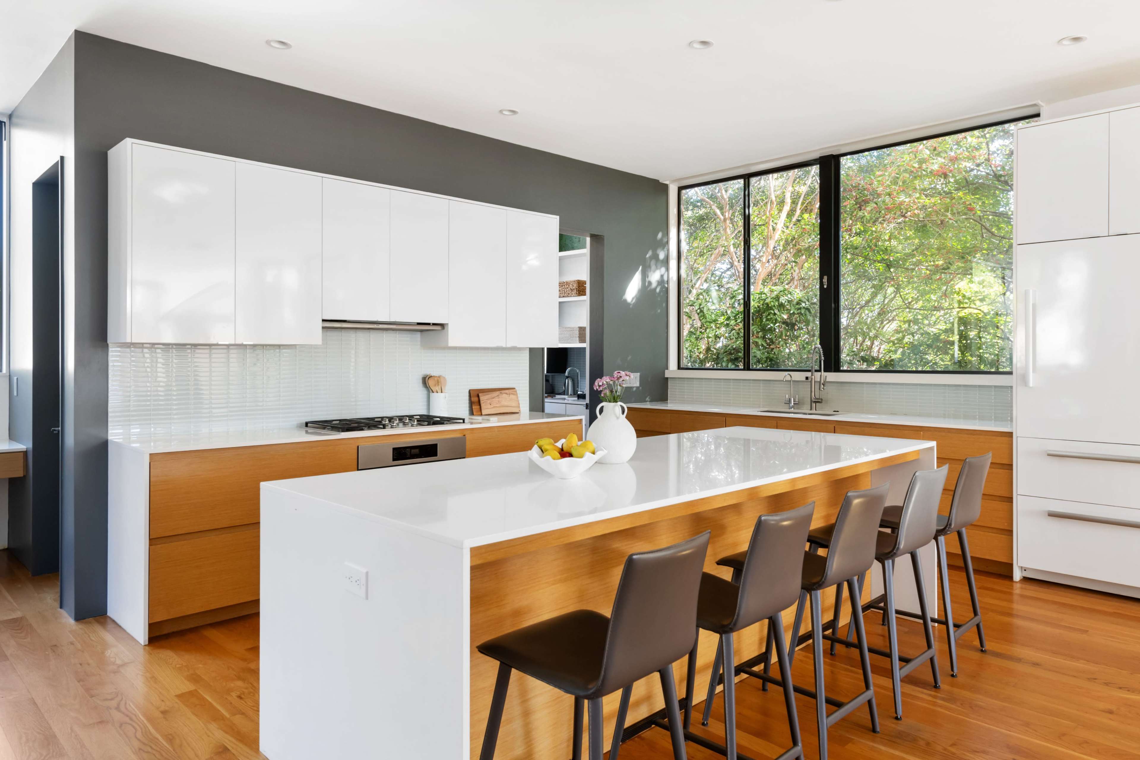 The kitchen features modern cabinetry, a large central island with seating, and large windows providing natural light and views of greenery.