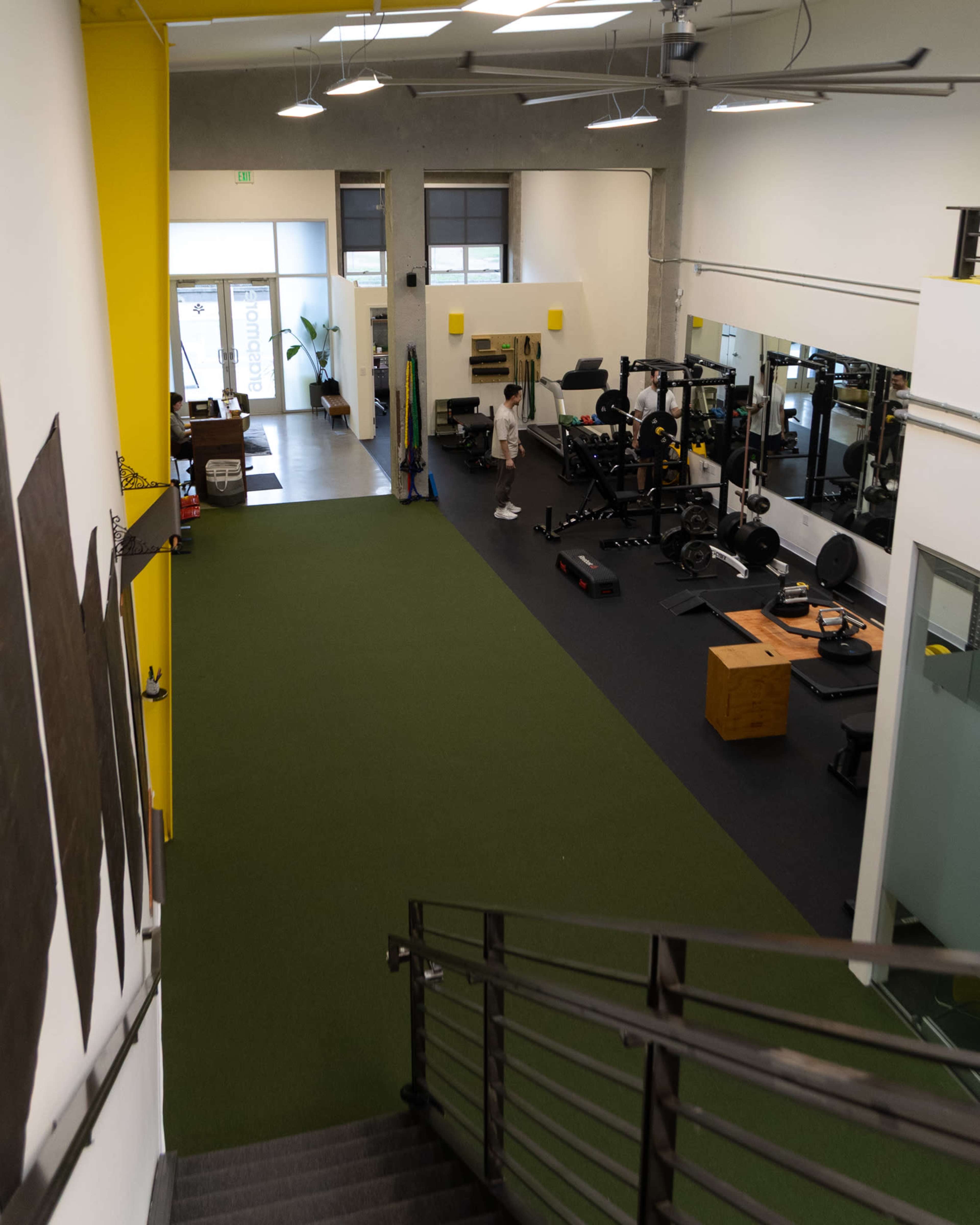 The image shows a spacious gym with weightlifting equipment, mirrors, and artificial grass flooring, viewed from a stairway.