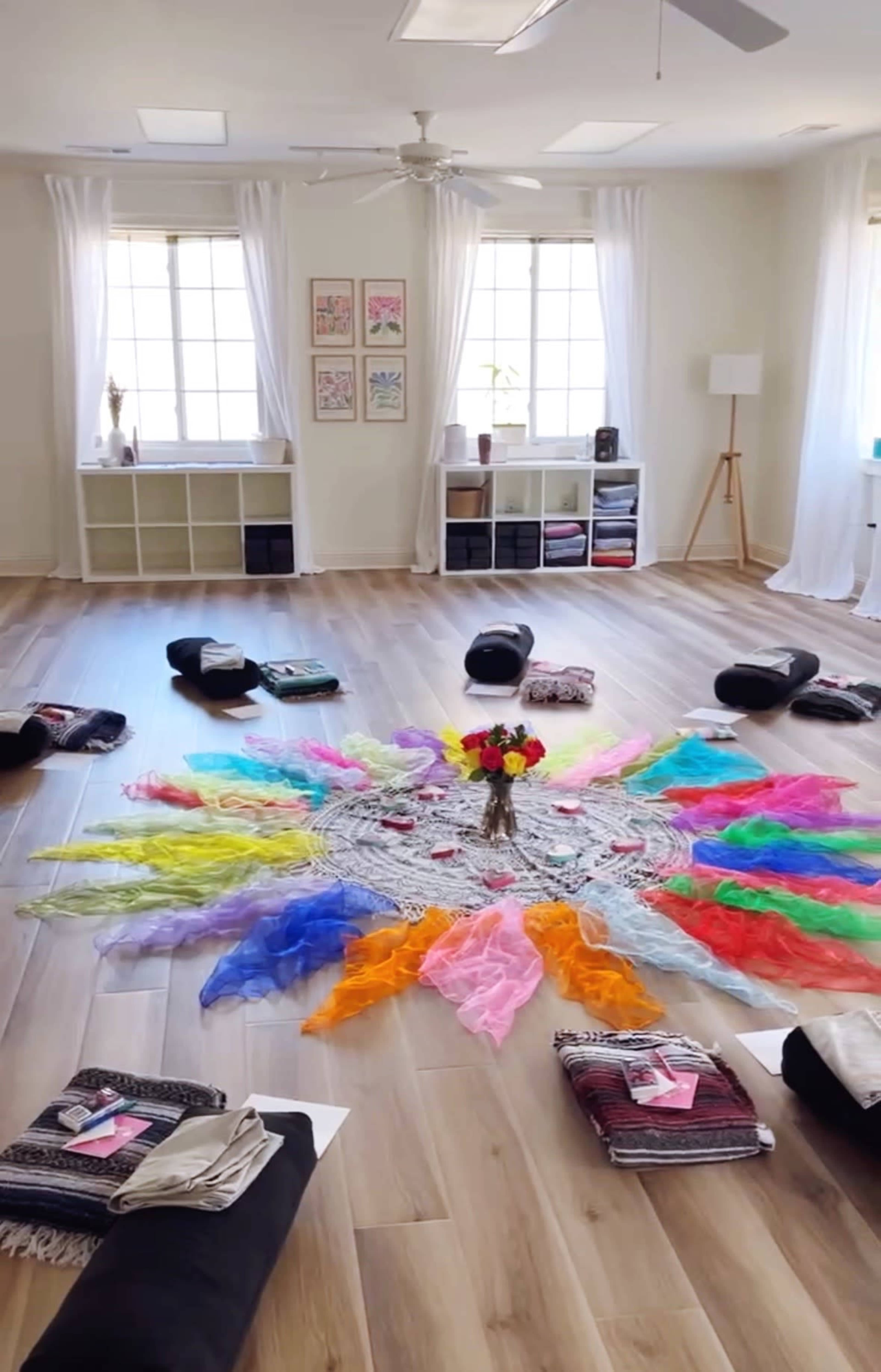 The image shows a spacious, well-lit room with colorful fabric draped in a circular pattern around a central flower arrangement on a lace doily, with yoga mats and folded textiles arranged around the perimeter.