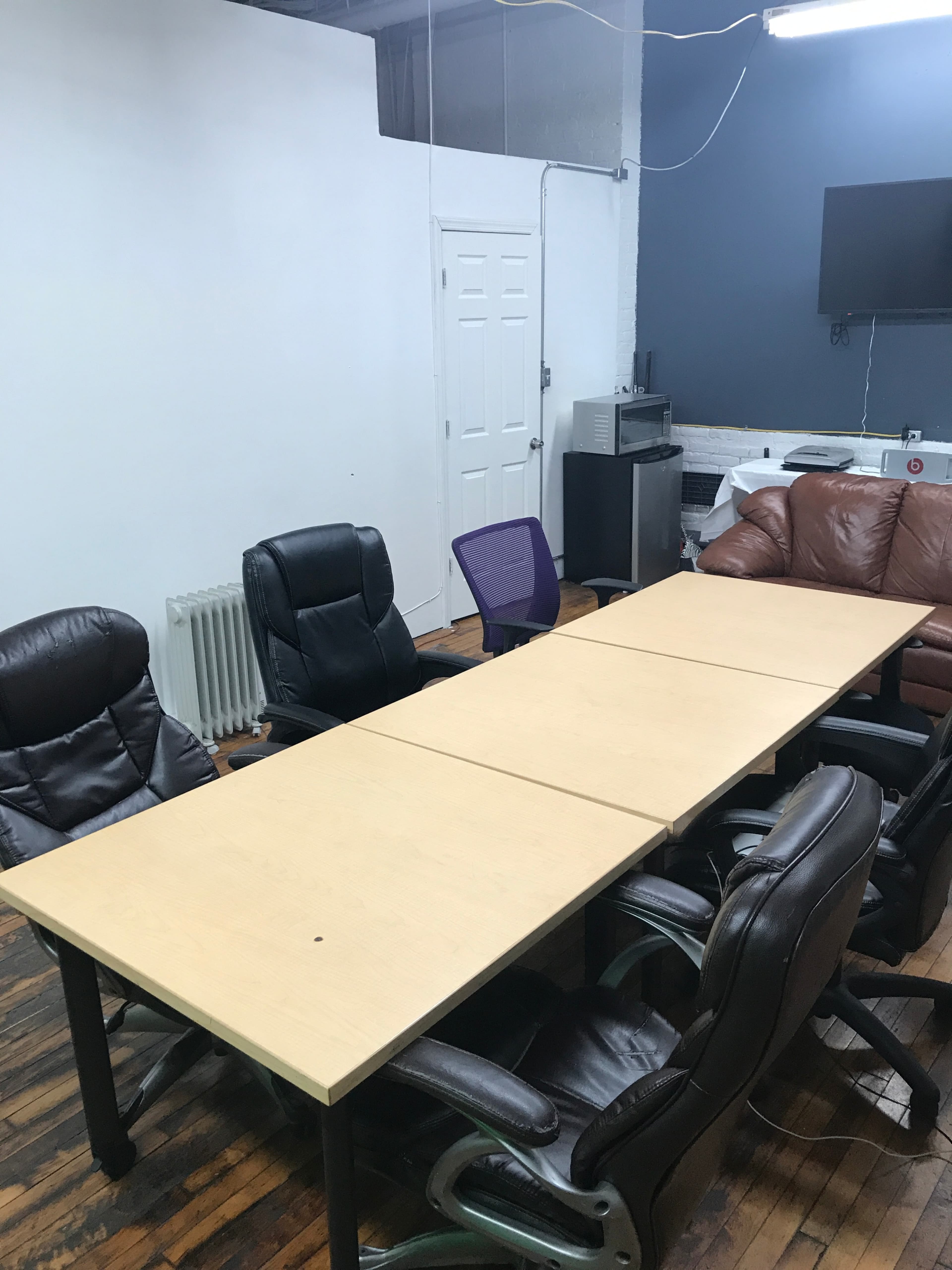 A long wooden table is surrounded by various office chairs in a meeting room with exposed flooring and a television on the wall.