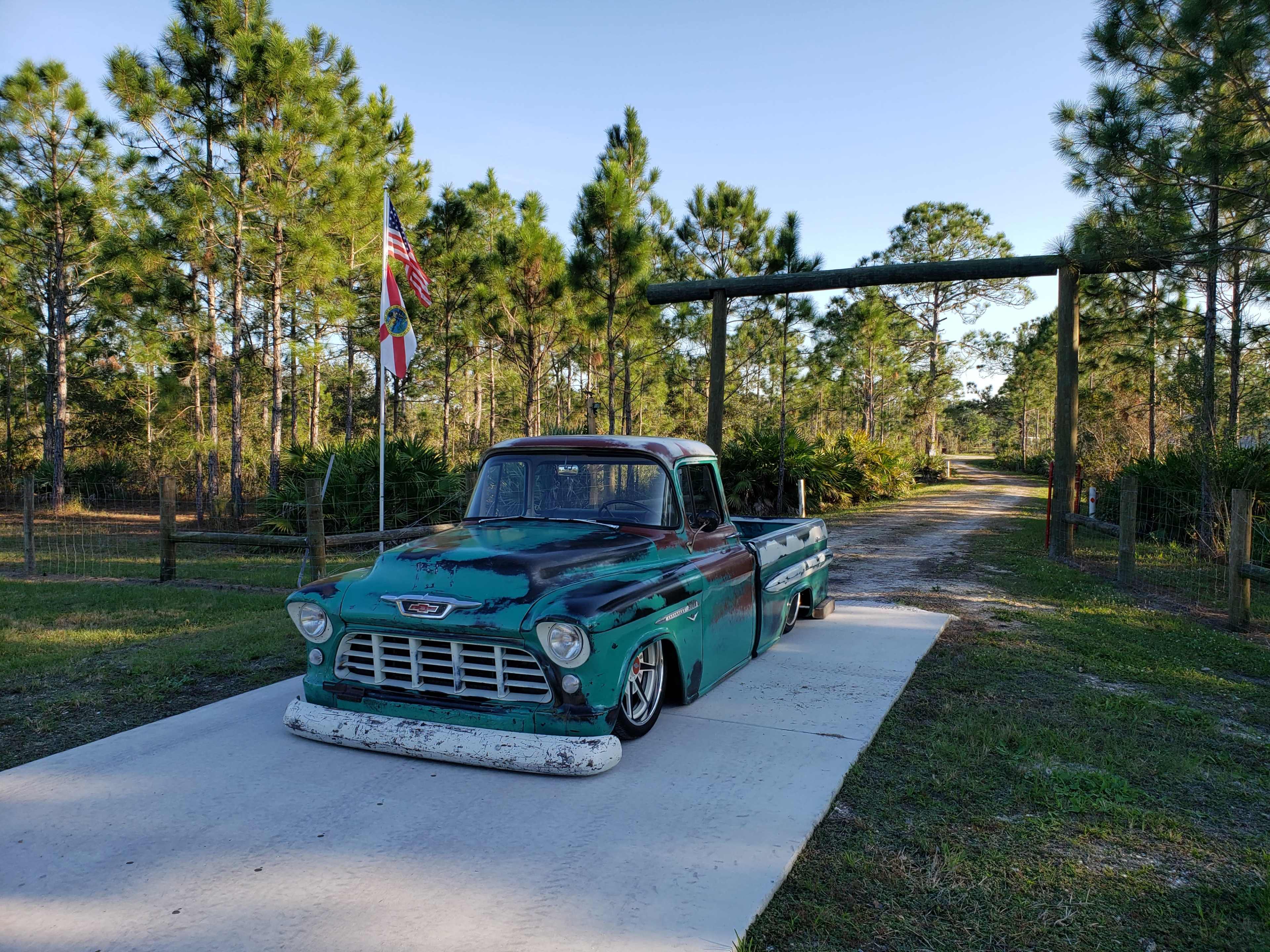 A vintage green pickup truck is parked on a concrete driveway leading to a tree-lined path with an American flag and a decorative signpost.