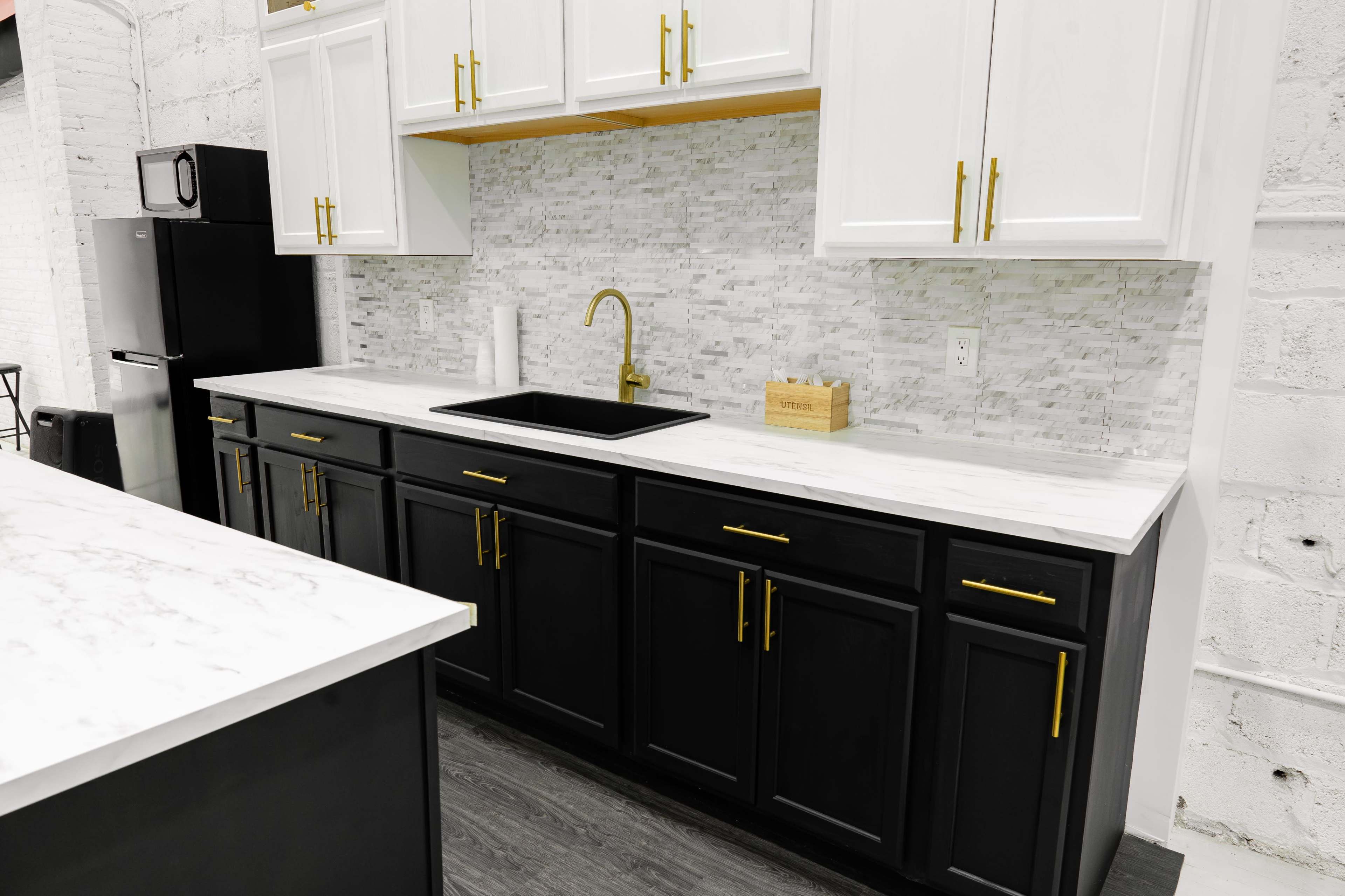 A modern kitchen with black lower cabinets, white upper cabinets, a marble countertop, and a gold faucet against a white and gray tiled backsplash.
