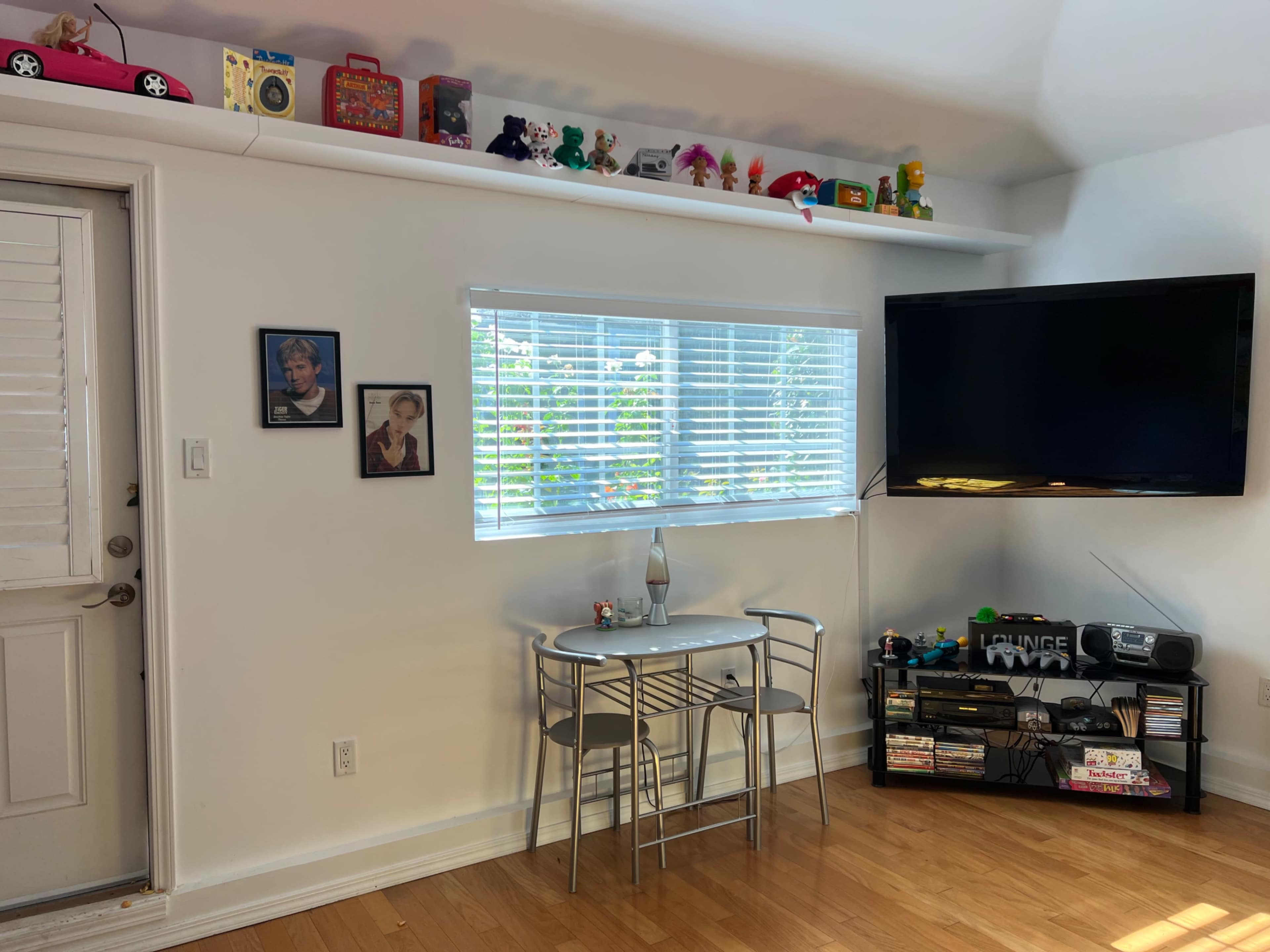 A small dining area with a round table and two chairs, next to a window, a wall-mounted television, and a shelf lined with colorful toys and decorations.