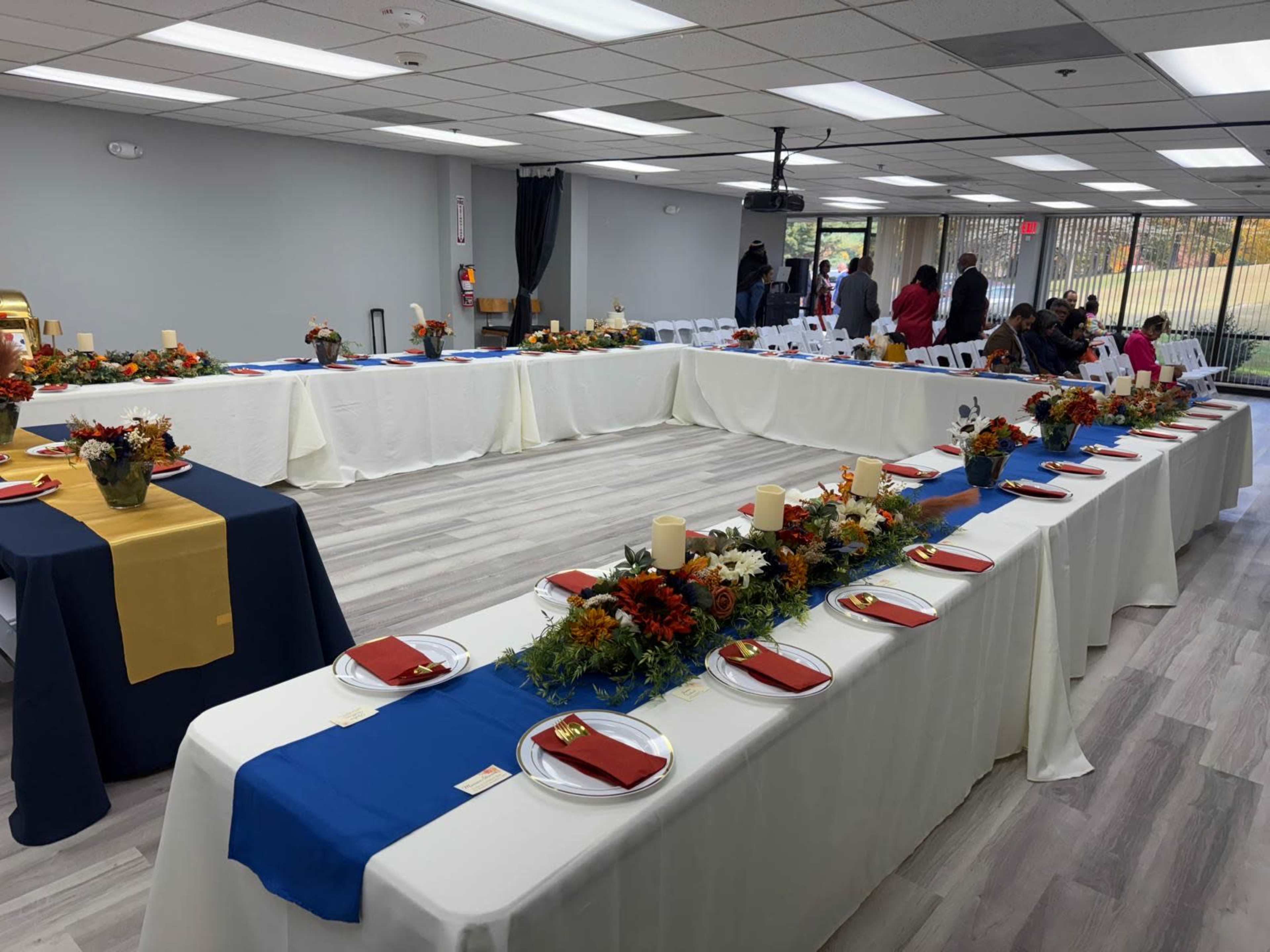 A large banquet room is set up with long tables covered in white and blue tablecloths, decorated with floral centerpieces and place settings, as people gather in the background.
