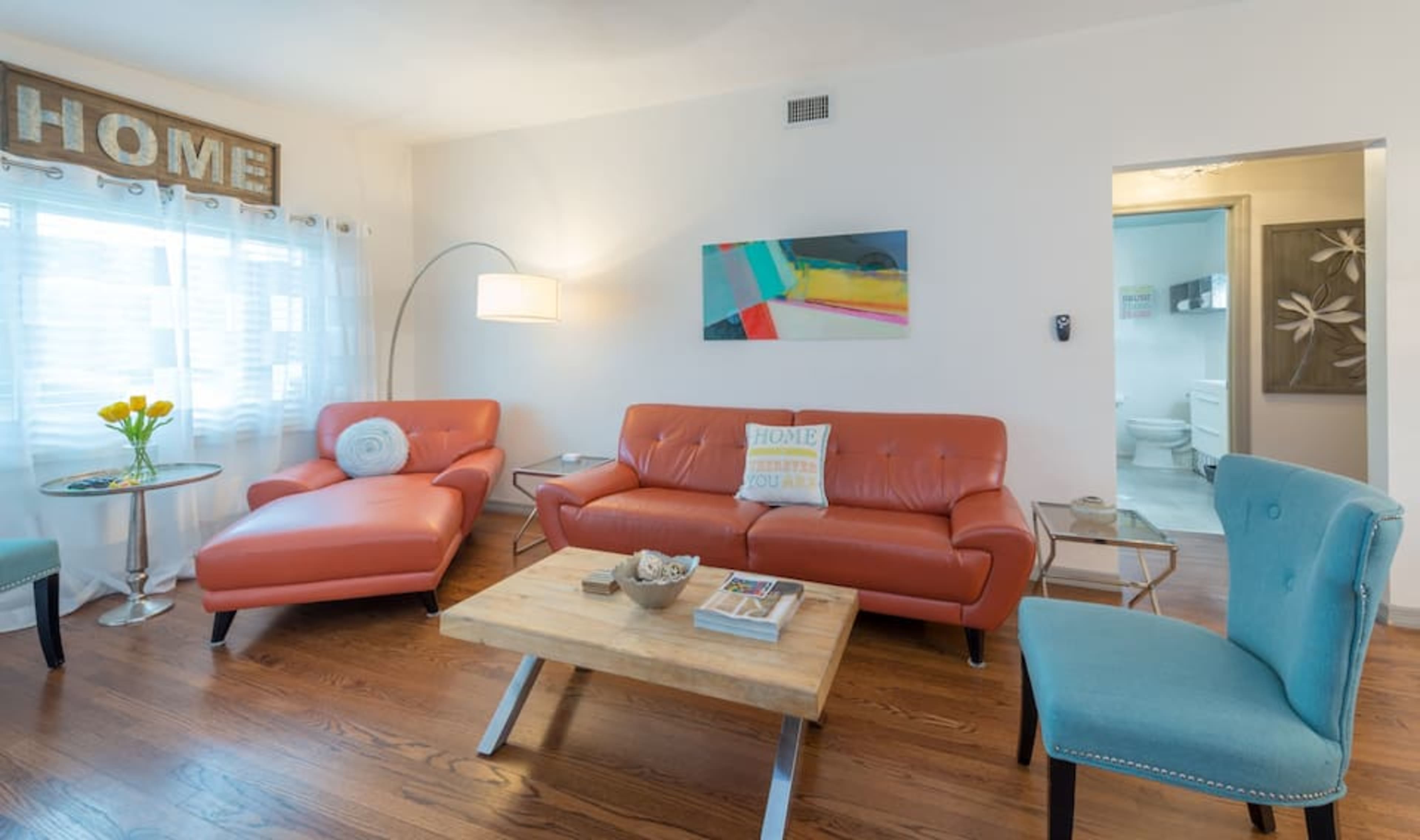 A bright living room featuring a coral sofa, a light blue accent chair, a wooden coffee table, and a decorative vase with yellow flowers.