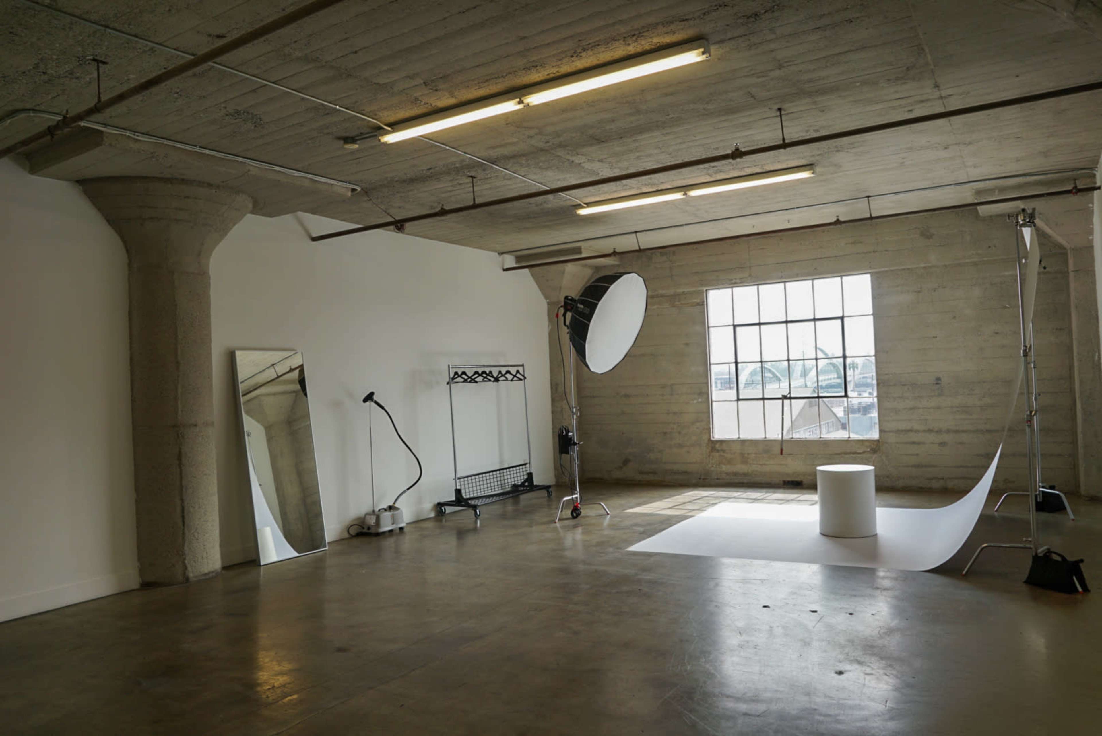The image shows a minimalist photography studio with a large window, a white backdrop, a circular prop, and various equipment including a reflector and a mirror.