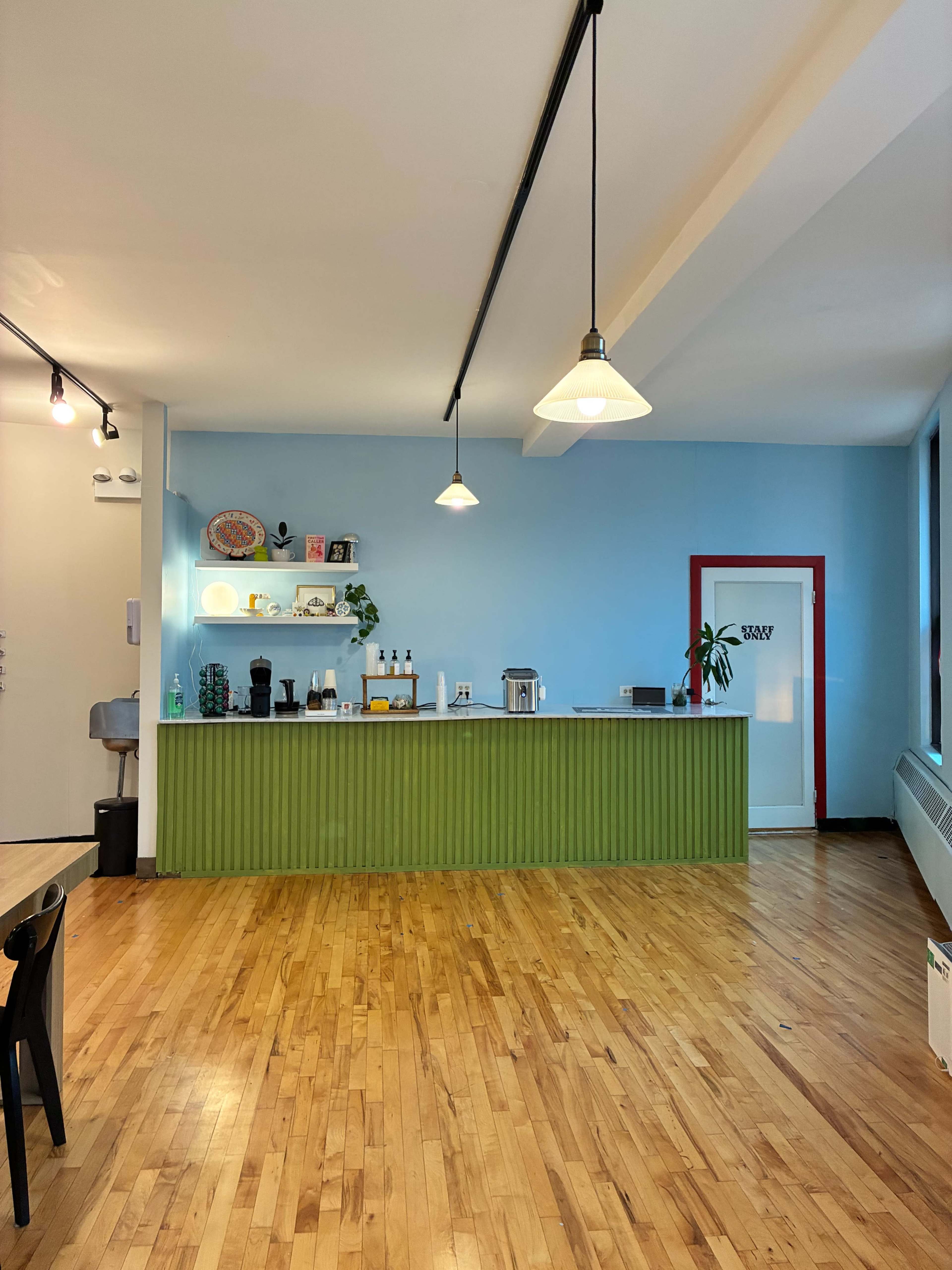 The image shows a minimalist café interior featuring a green counter, light blue walls, wooden flooring, and a doorway with a red frame.