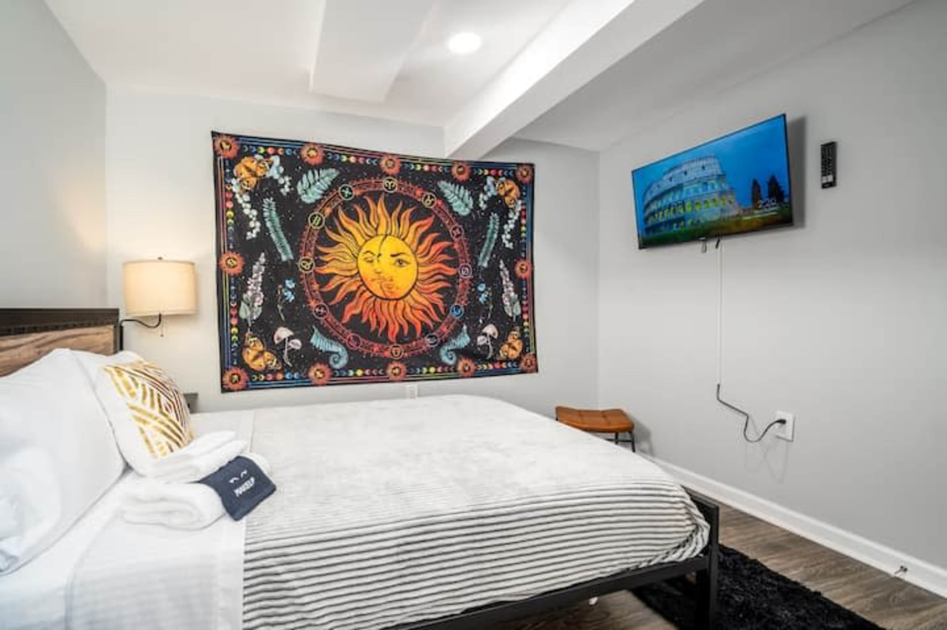 A bedroom with a bed, a tapestry featuring a sun design on the wall, and a television mounted nearby.