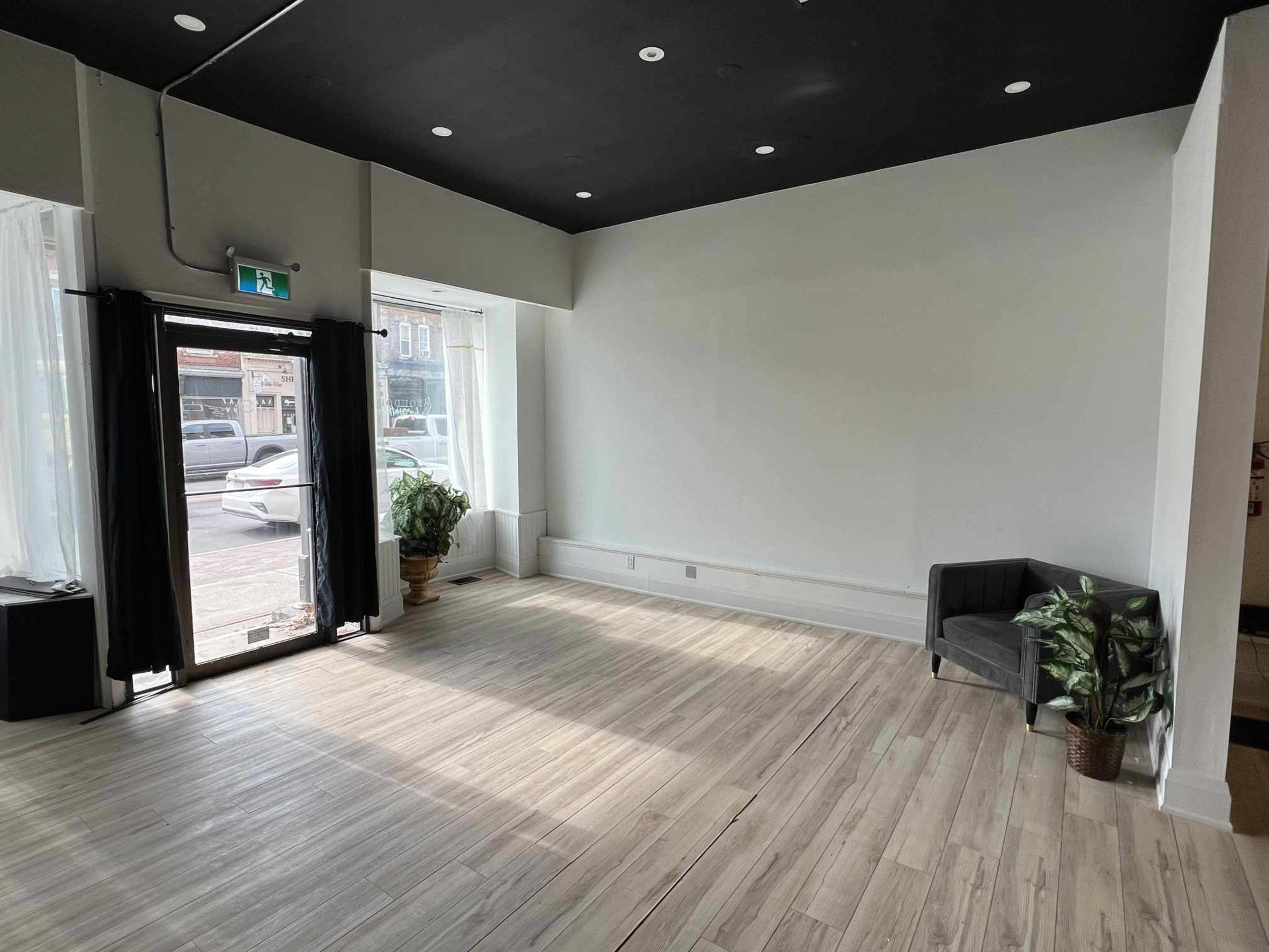 An empty room features hardwood flooring, a large window with sheer curtains, a potted plant, and a gray sofa against the wall.