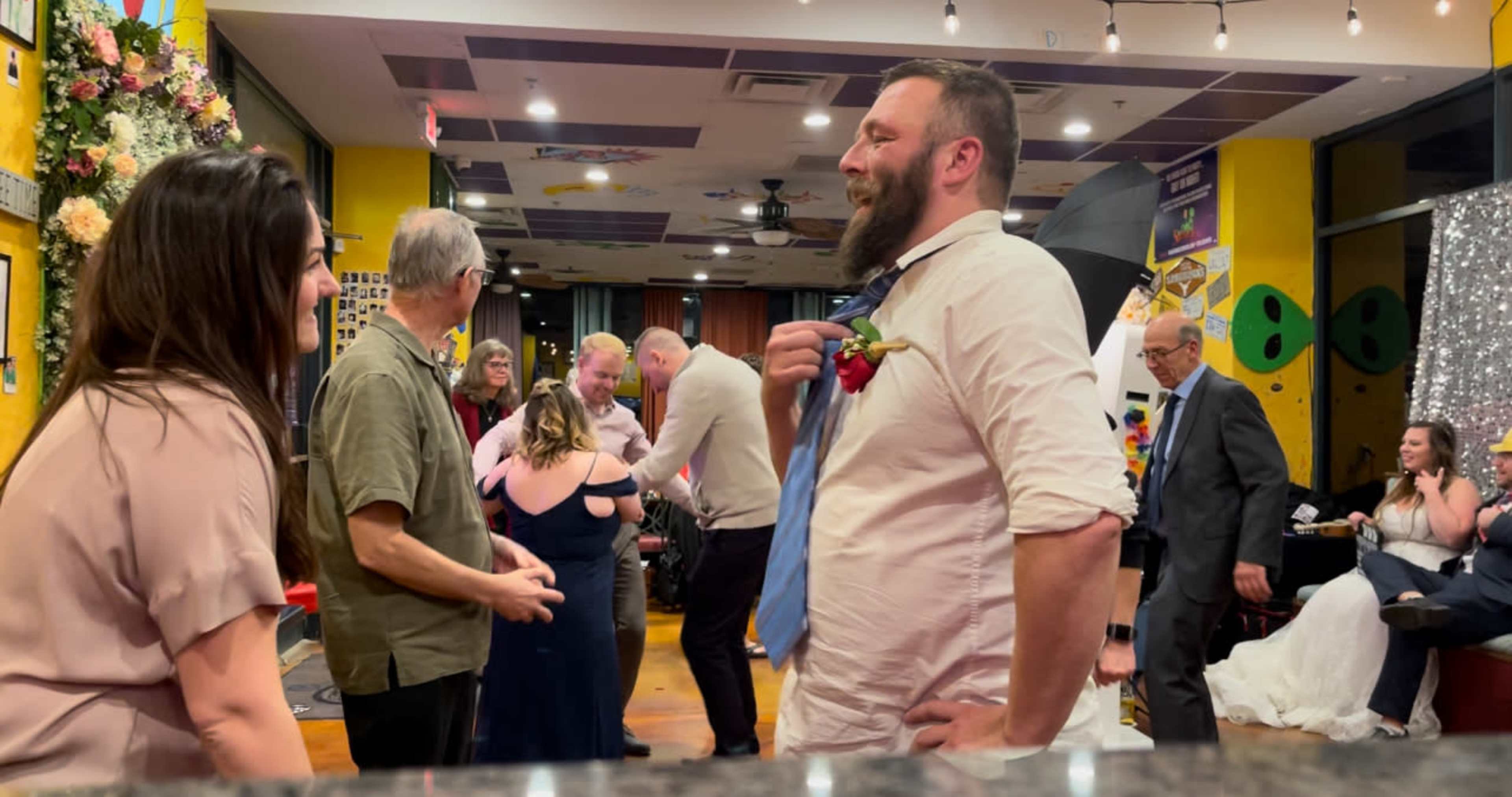 A group of people socialize and dance at a festive indoor gathering, with floral decorations and a few individuals dressed in formal attire.