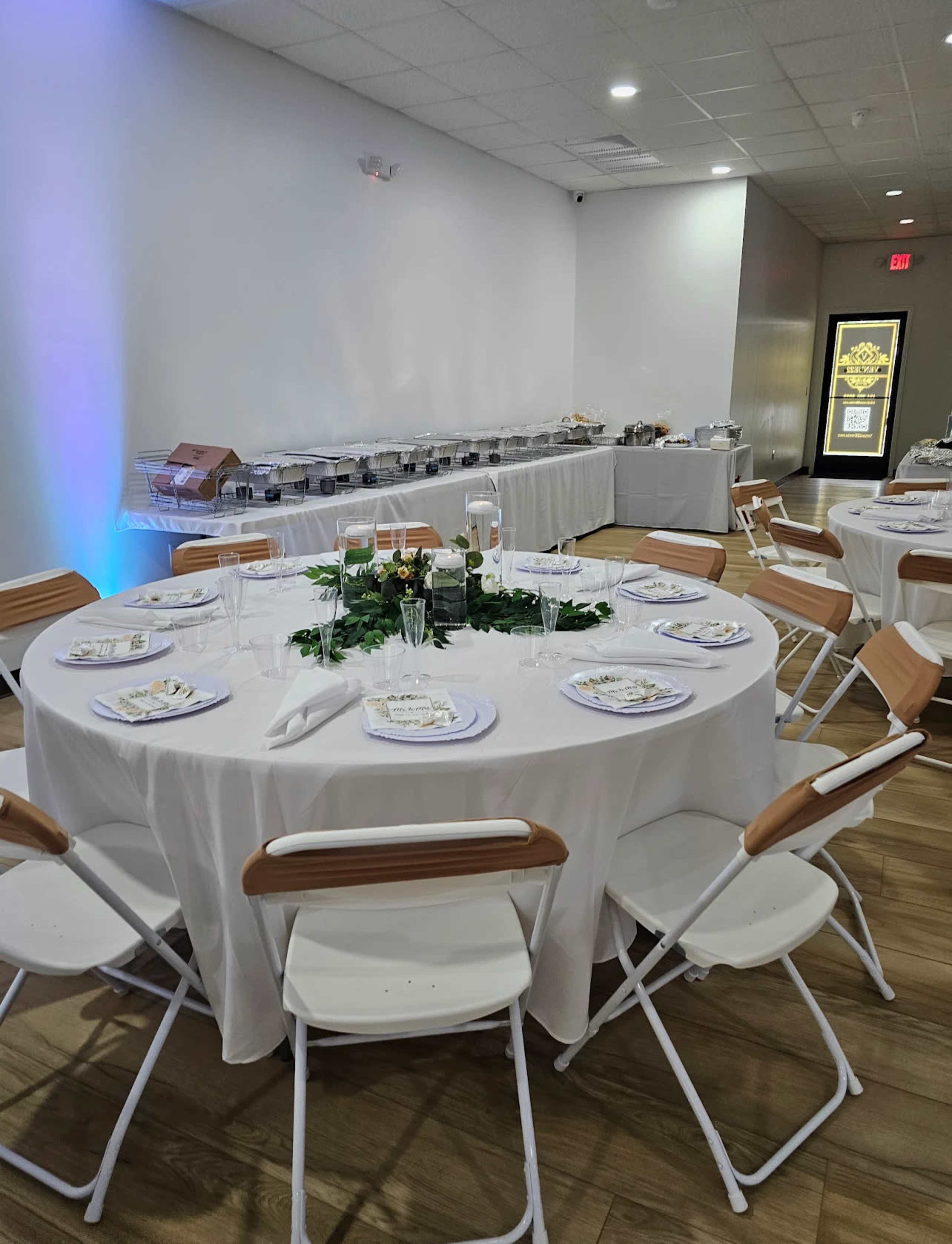 A dining arrangement features round tables with white tablecloths, set with plates and glasses, in a spacious room with a buffet setup in the background.