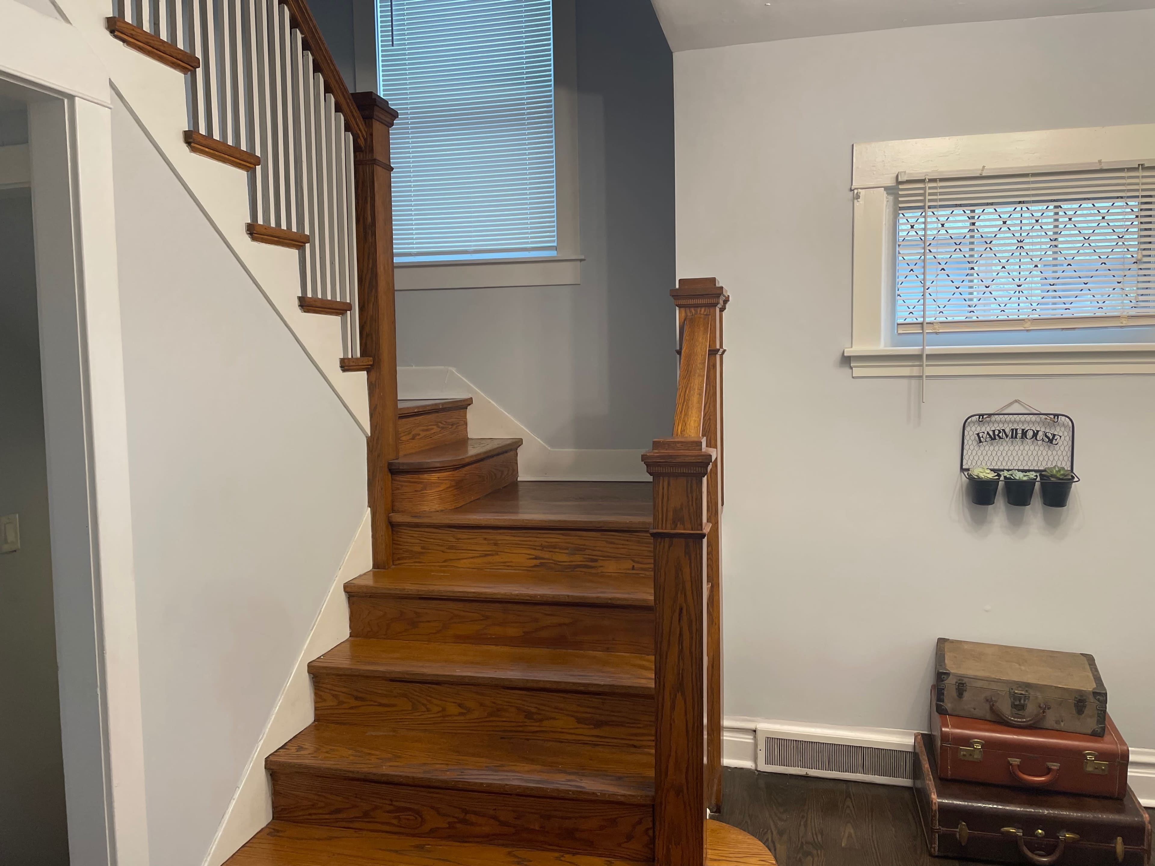 A wooden staircase with a banister leads upward, next to a wall with a window and a small table holding vintage suitcases.