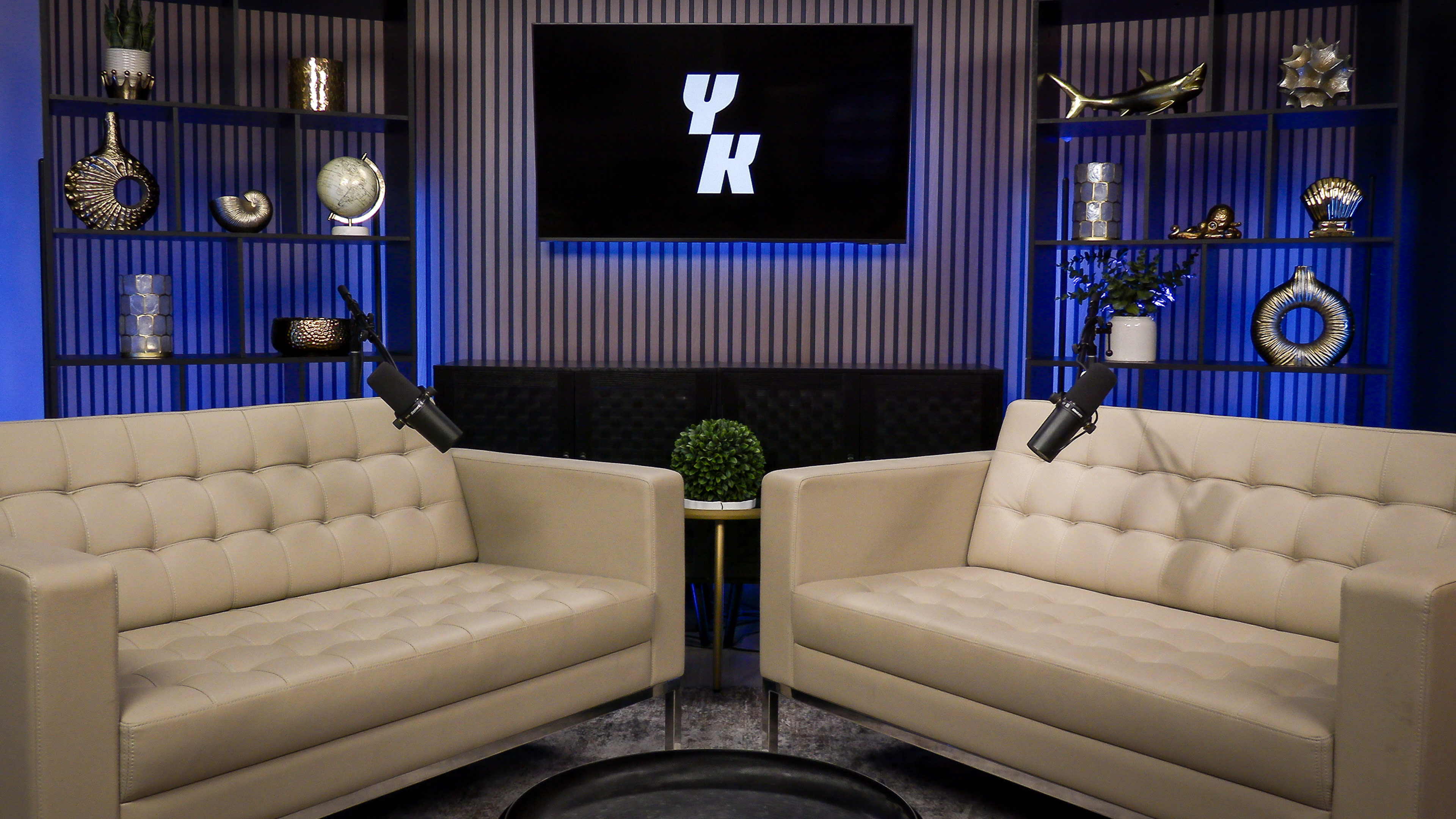 The image shows a modern studio setup with two white tufted sofas facing each other, a small black table in between, and decorative shelves in the background featuring various ornamental objects and a wall-mounted screen displaying the letters "YK."