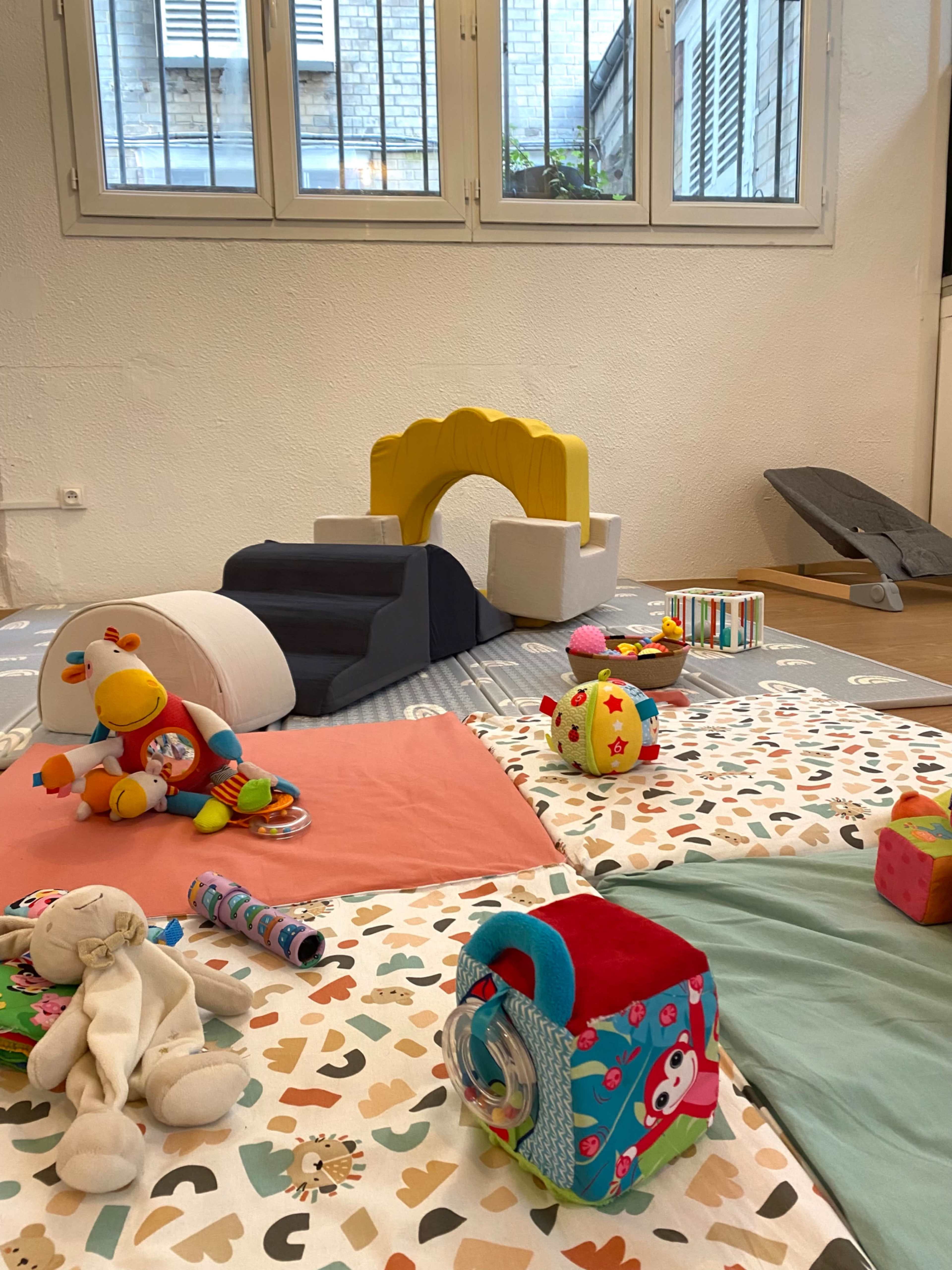 The image shows a playroom with various toys scattered across colorful mats, including plush toys, building blocks, and a small slide-like structure.