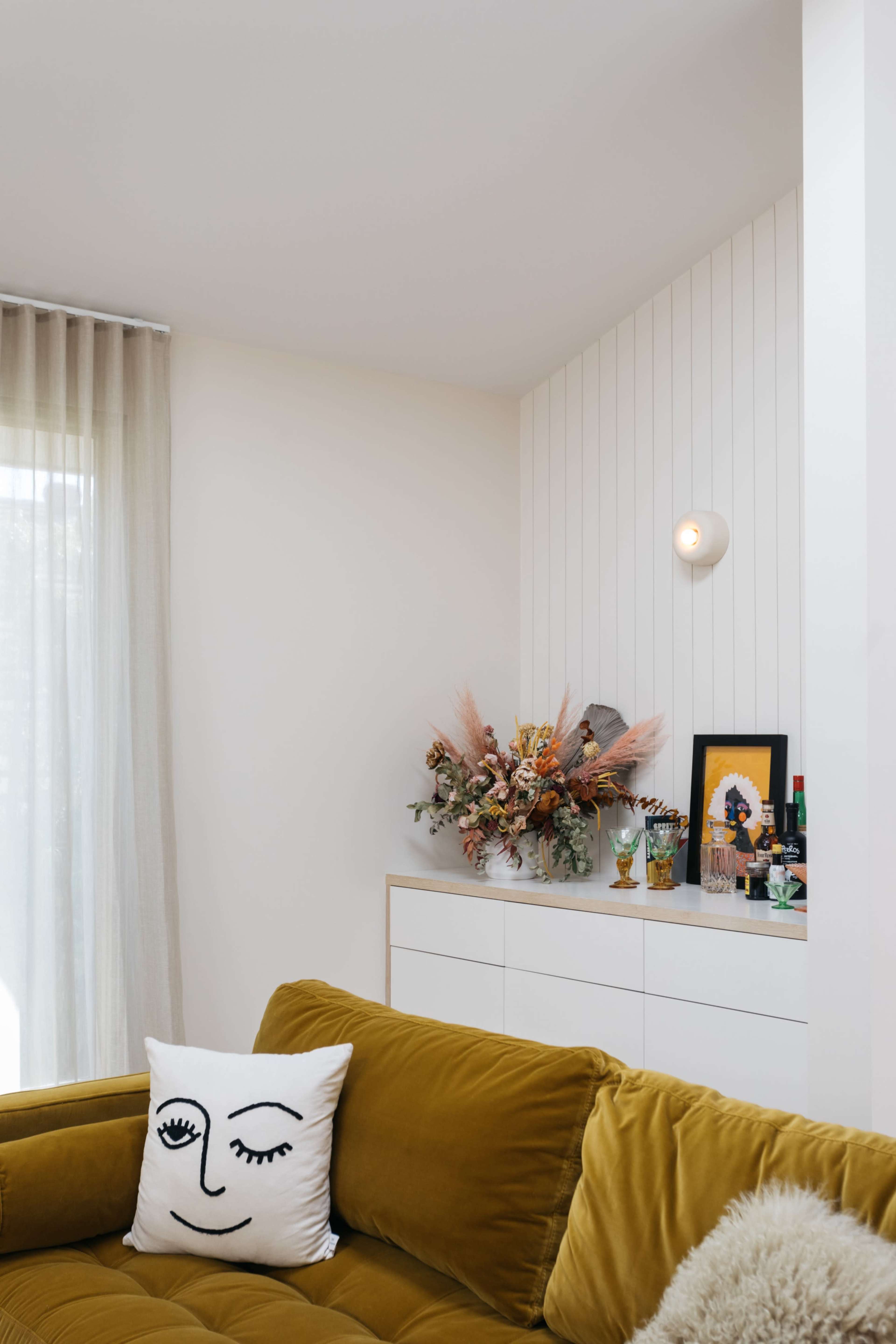 A cozy living room features a mustard-colored sofa, a decorative pillow with a face design, and a shelf adorned with a bouquet and framed artwork.