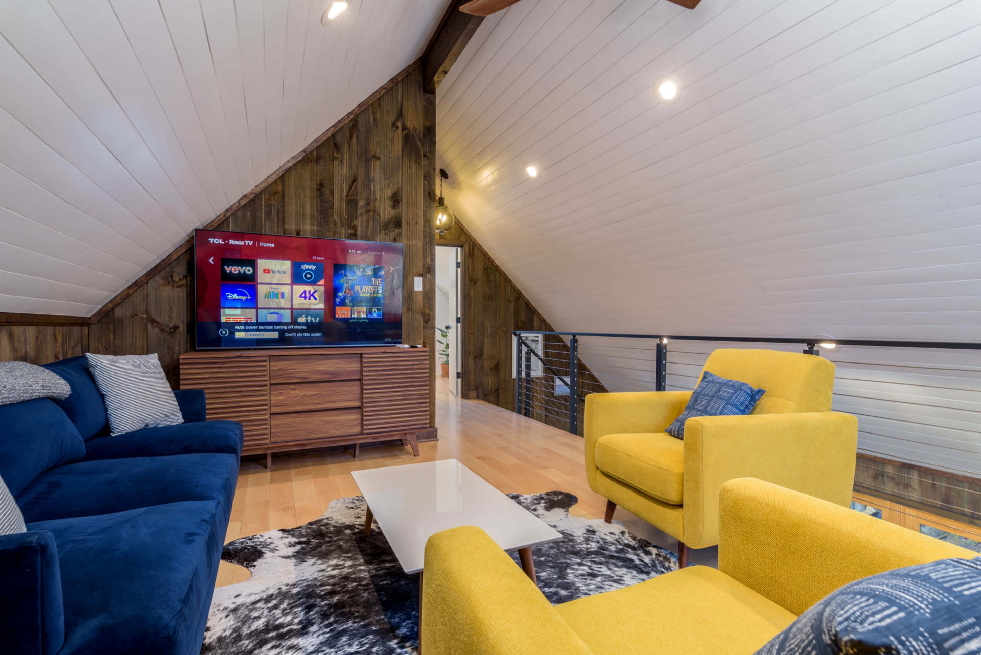 A cozy loft living area with a blue sectional sofa, yellow armchairs, and a wall-mounted television displaying various streaming options.