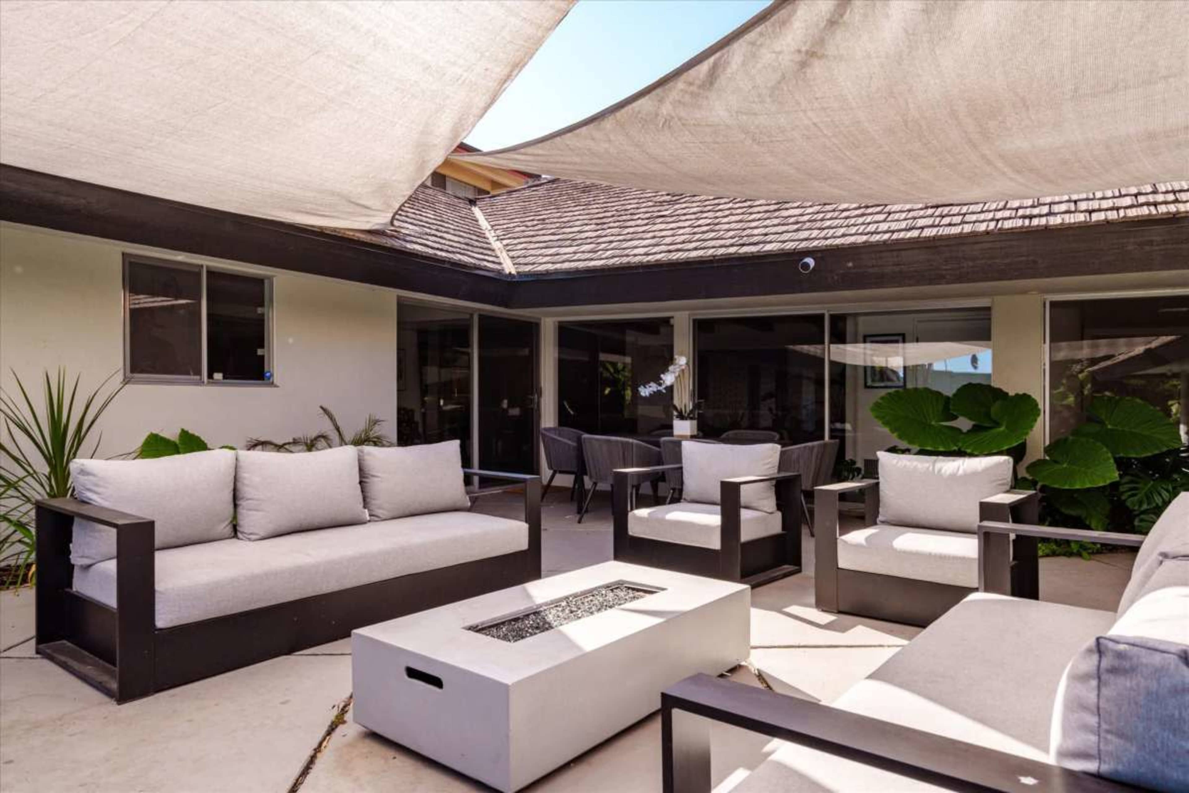 The image shows a patio area with several gray outdoor sofas and a fire pit, surrounded by plants and shaded by large fabric canopies.