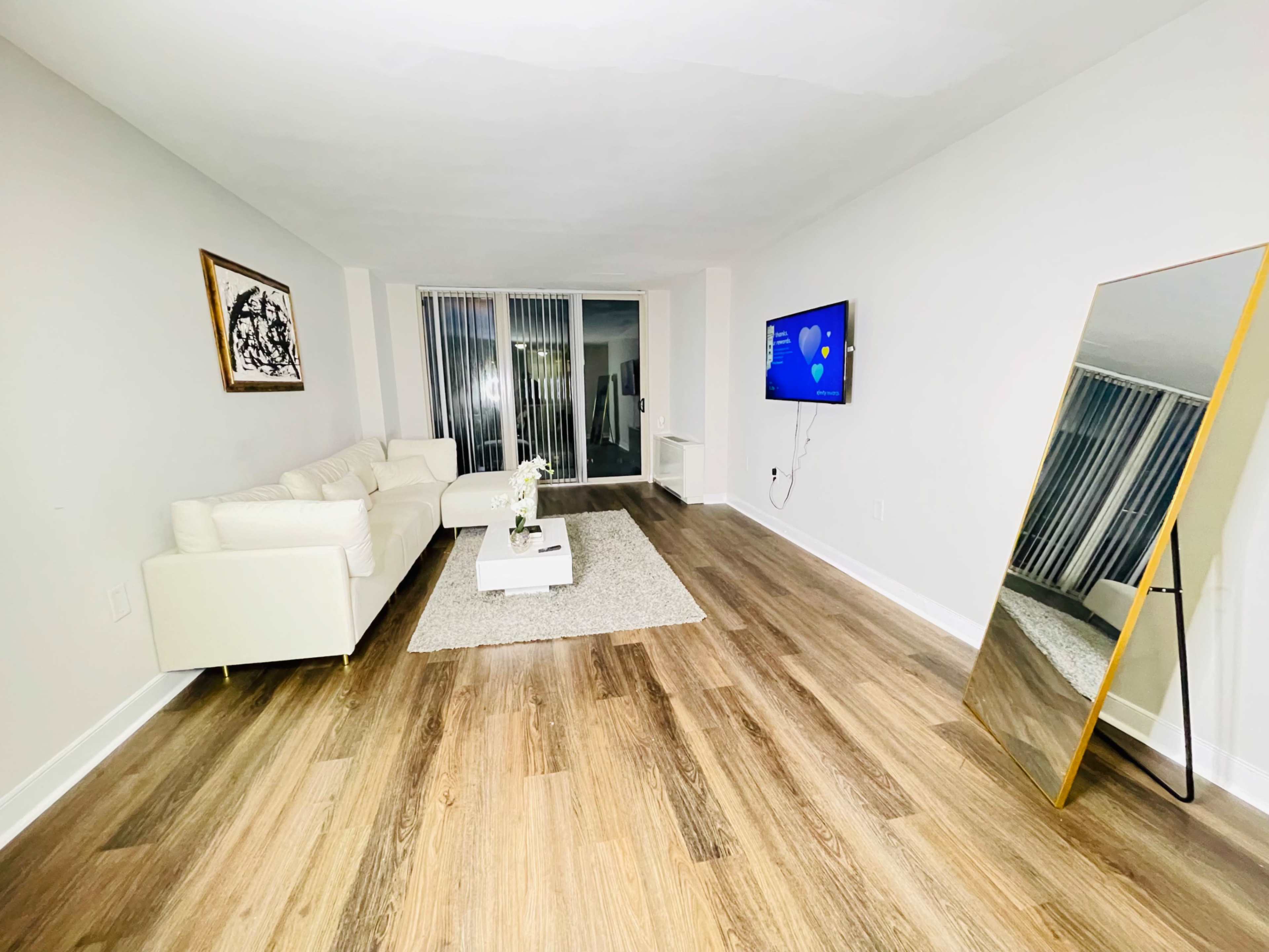 A modern, minimalist living room features a white sofa, a large mirror, a flat-screen TV, and hardwood flooring.