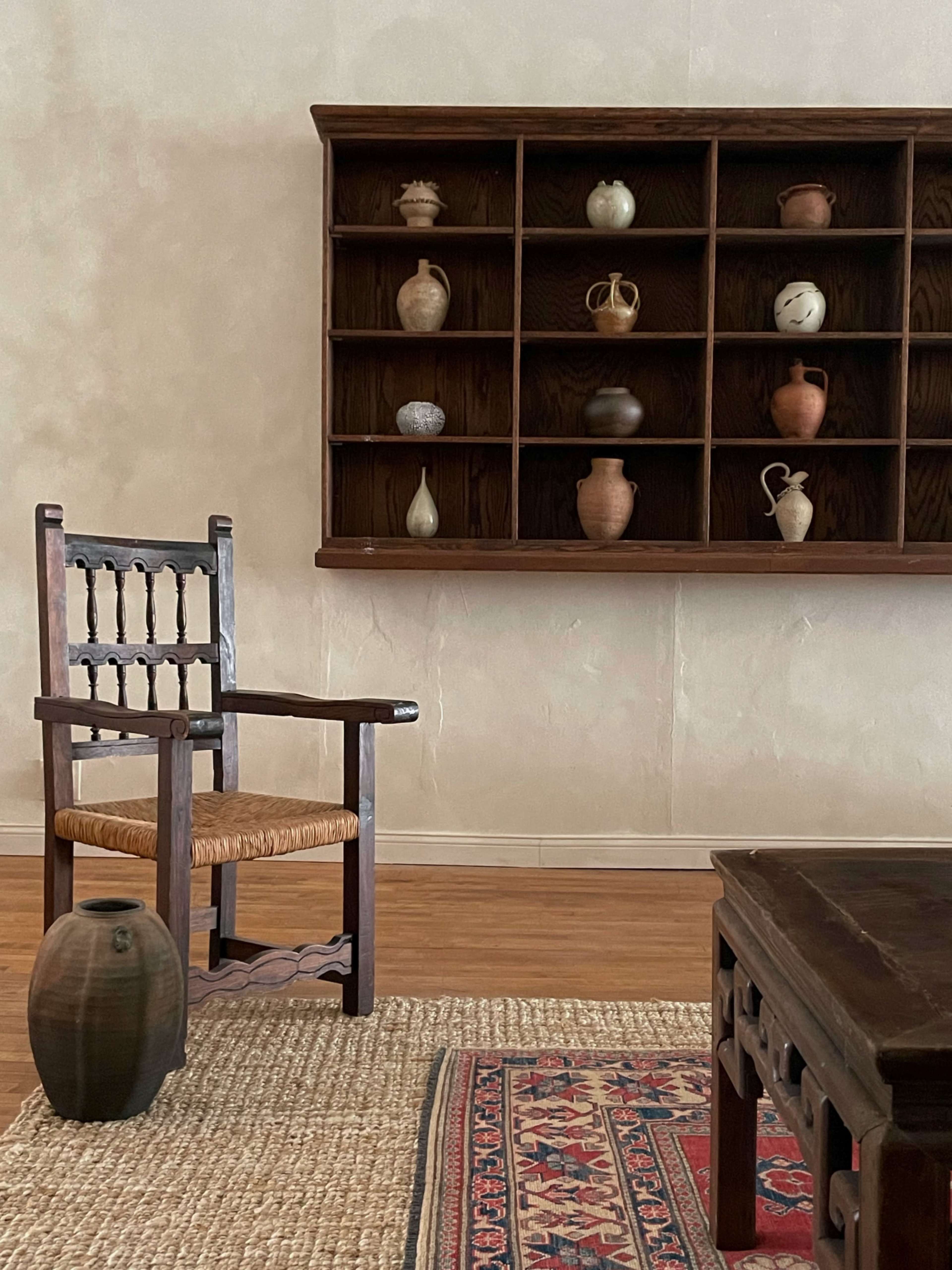 A simple room with a wooden chair, a low table, an earthenware pot, and a wall shelf displaying various pottery items.