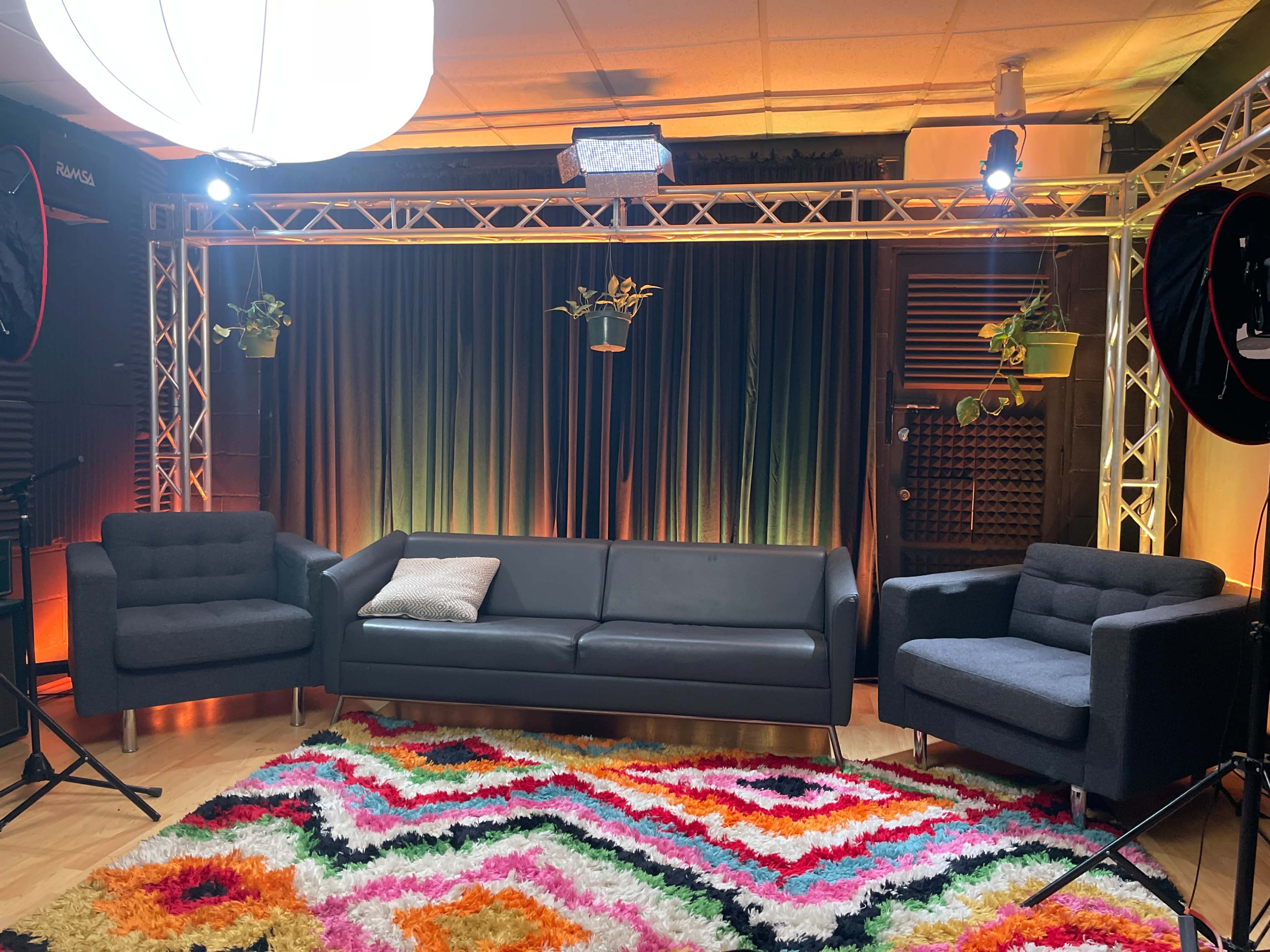 The image shows a brightly lit studio setup with a black couch, two armchairs, and a colorful rug on the wooden floor, surrounded by curtains and various lighting equipment.
