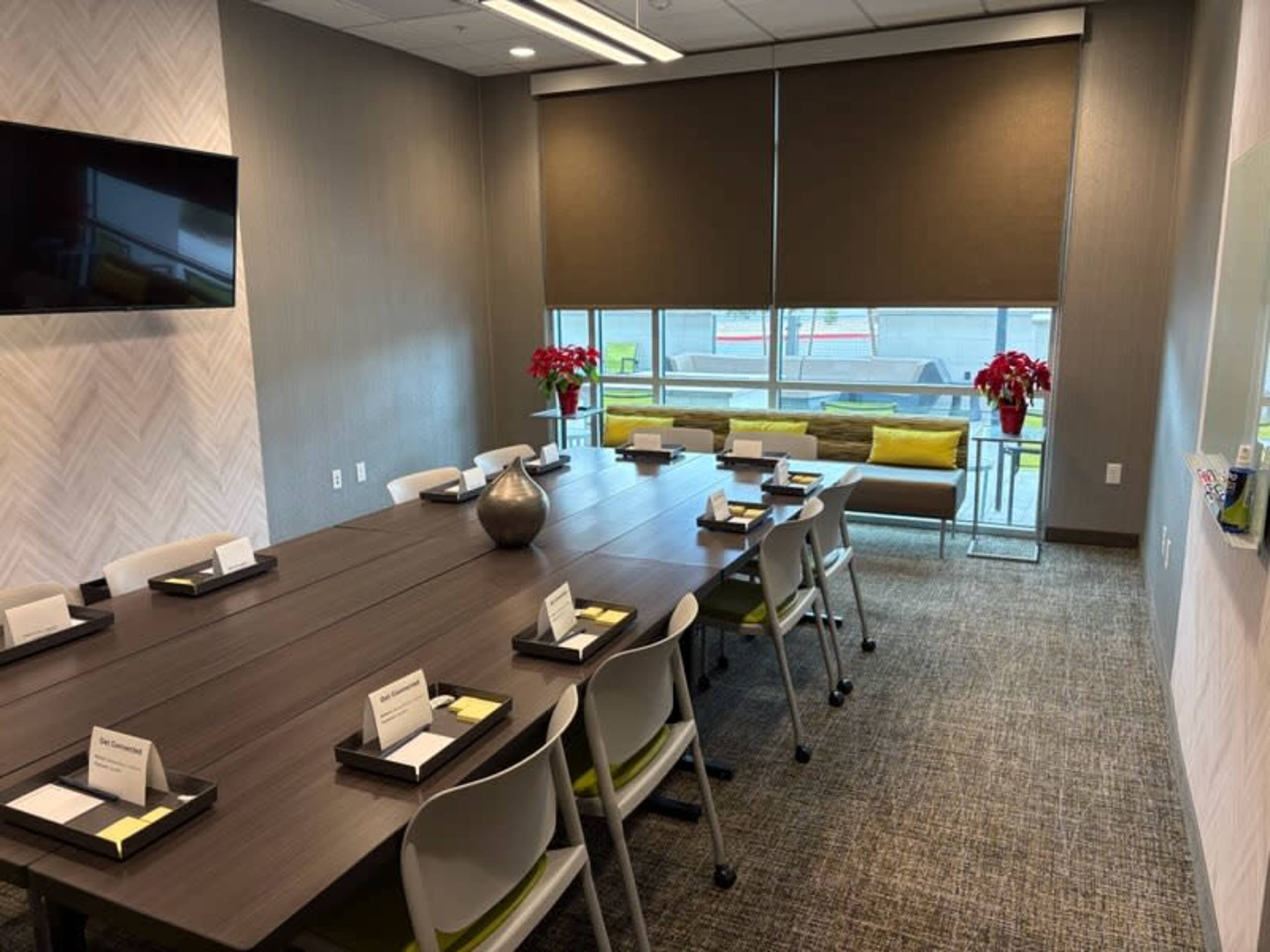 A conference room features a long wooden table surrounded by chairs, with floral arrangements on the table and a sofa against the window.