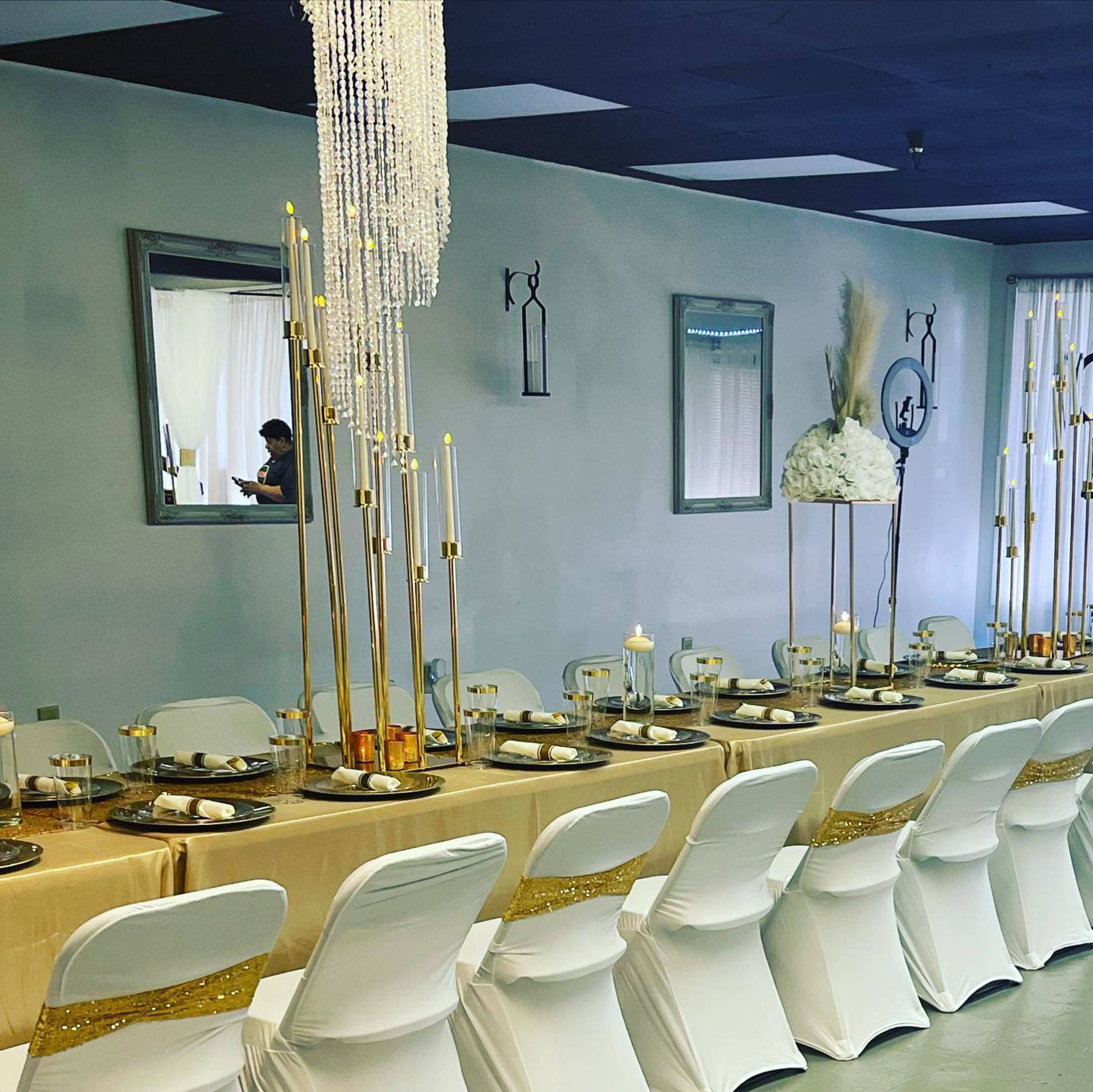 A long banquet table is elegantly set with gold and white decor, complemented by tall candle holders and a floral centerpiece, in a well-lit event space.