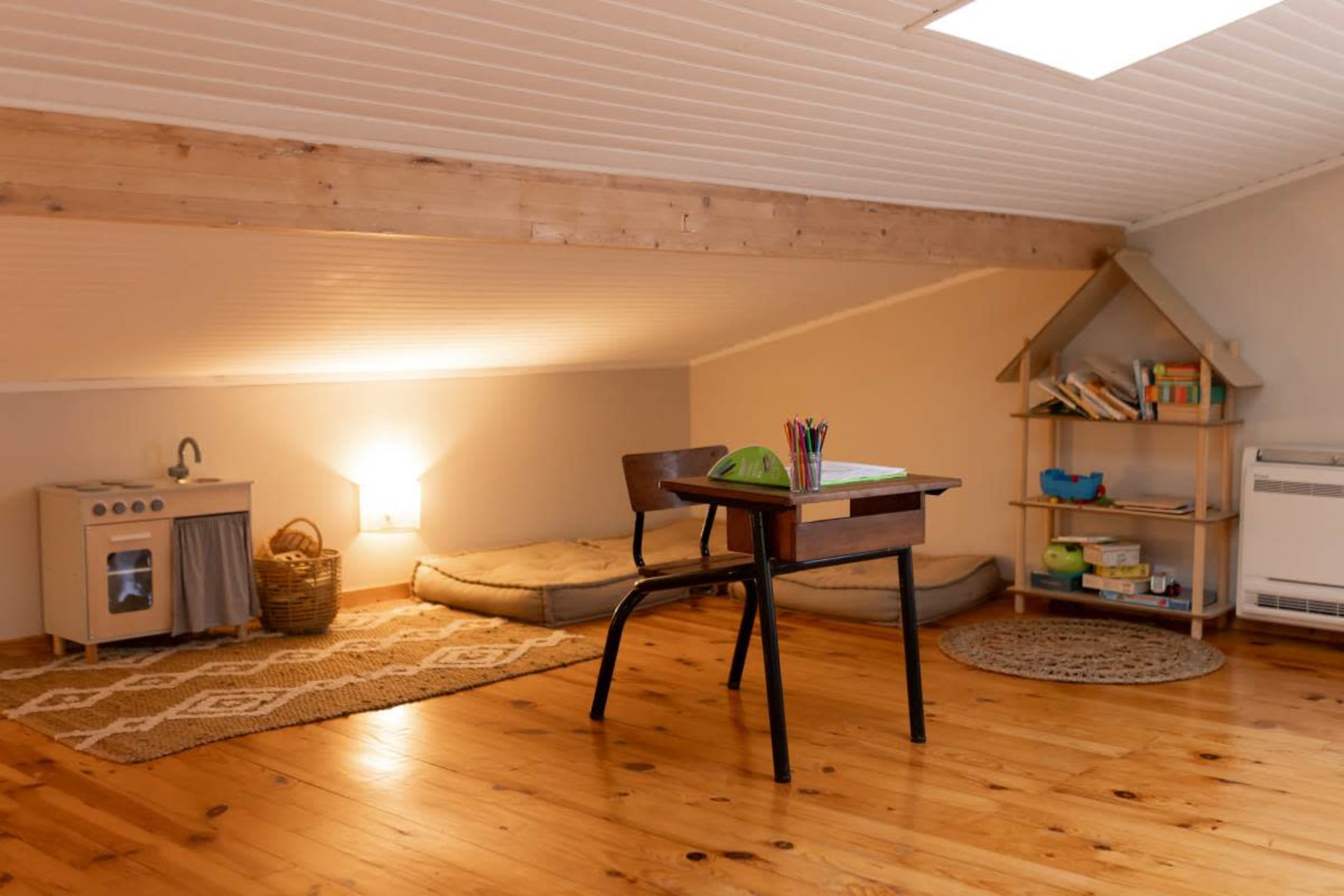 The image shows a cozy attic space with wooden flooring, a small desk with stationery, a play kitchen, and a reading nook with cushions and bookshelves.